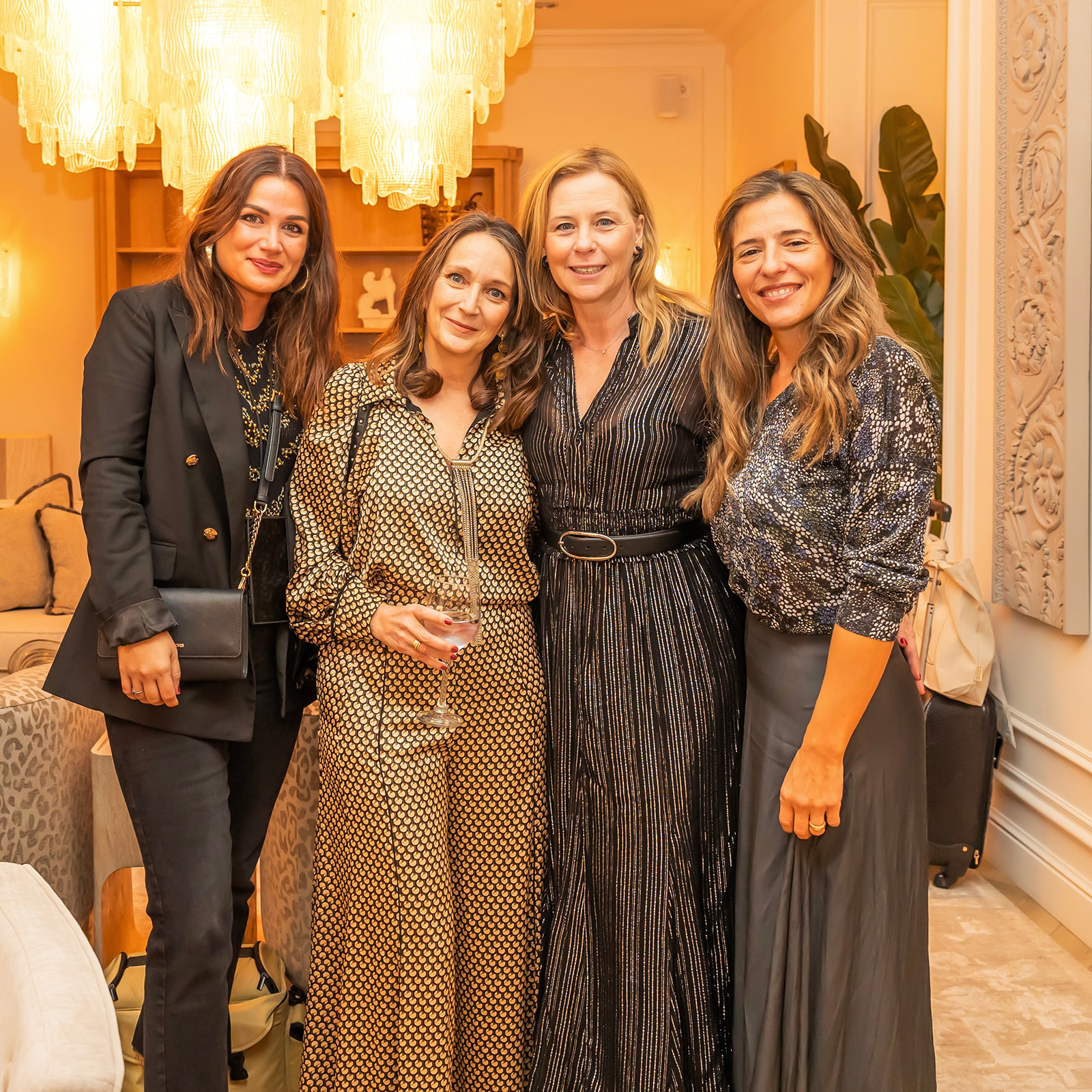 Cuatro mujeres sonrientes posando juntas en un elegante interior con una lámpara de araña. Visten ropa formal y de fiesta, incluyendo vestidos largos y un traje negro.