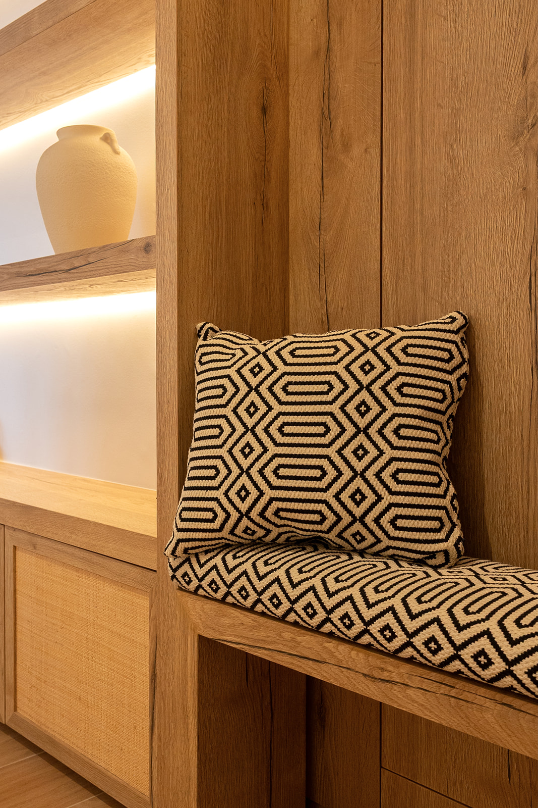 Estantería de madera con luz ambiental, jarrón decorativo en un estante superior y asiento acogedor con cojines estampados geométricos en tonos beige y negro