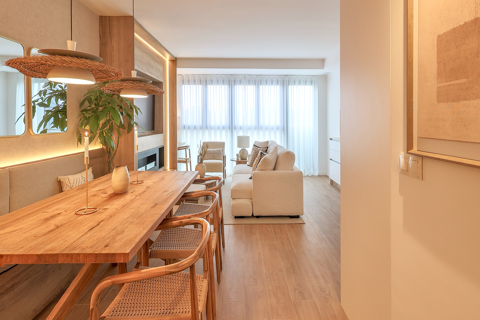 Sala de estar y comedor de estilo escandinavo con mesa de madera, lámparas de ratán, sofá blanco, y grandes ventanales. Decoración en tonos cálidos y naturales con plantas y elementos de madera.