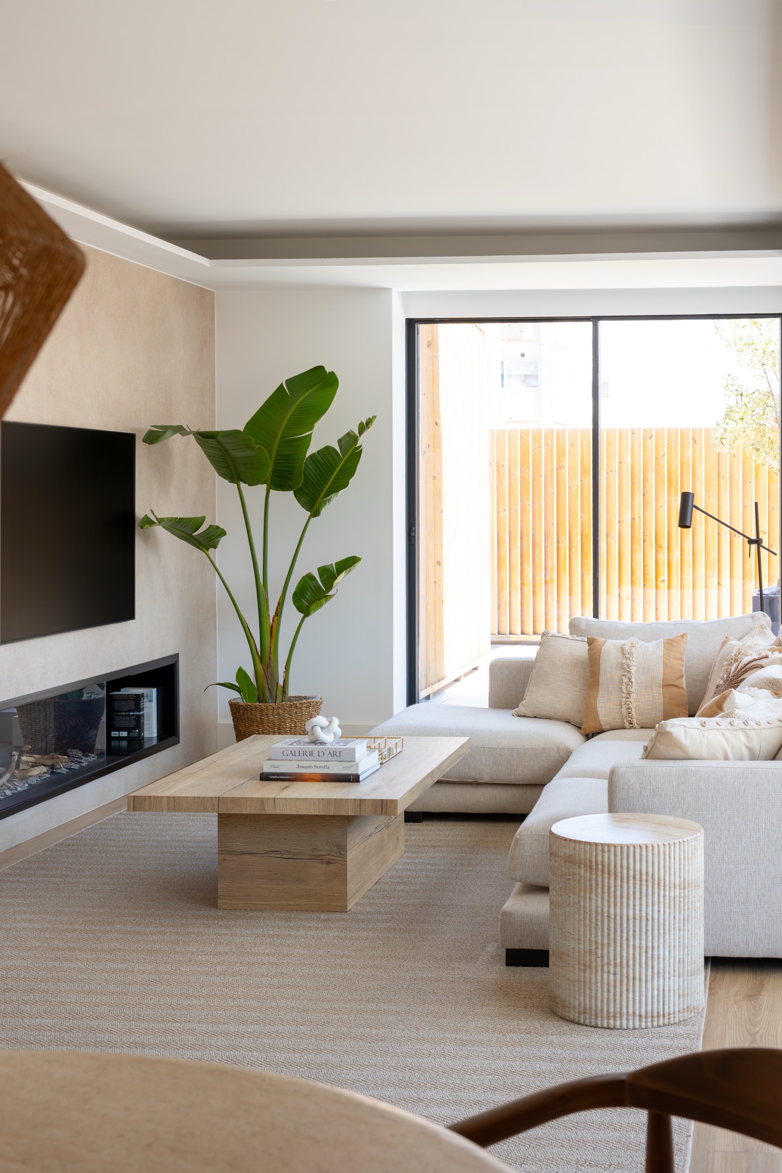 Sala de estar moderna y minimalista con sofá beige, mesa de centro de madera, planta grande en maceta, televisor en la pared y chimenea. Ventanal con vista a un jardín con valla de madera.