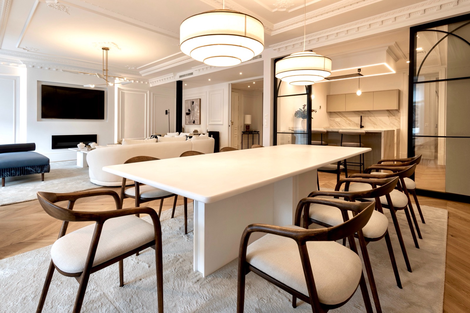 Elegante sala de estar y comedor con diseño interior moderno. Mesa de comedor blanca con sillas de madera, lámparas colgantes circulares, sofá blanco, televisor de pantalla plana y cocina abierta con acabados de mármol.