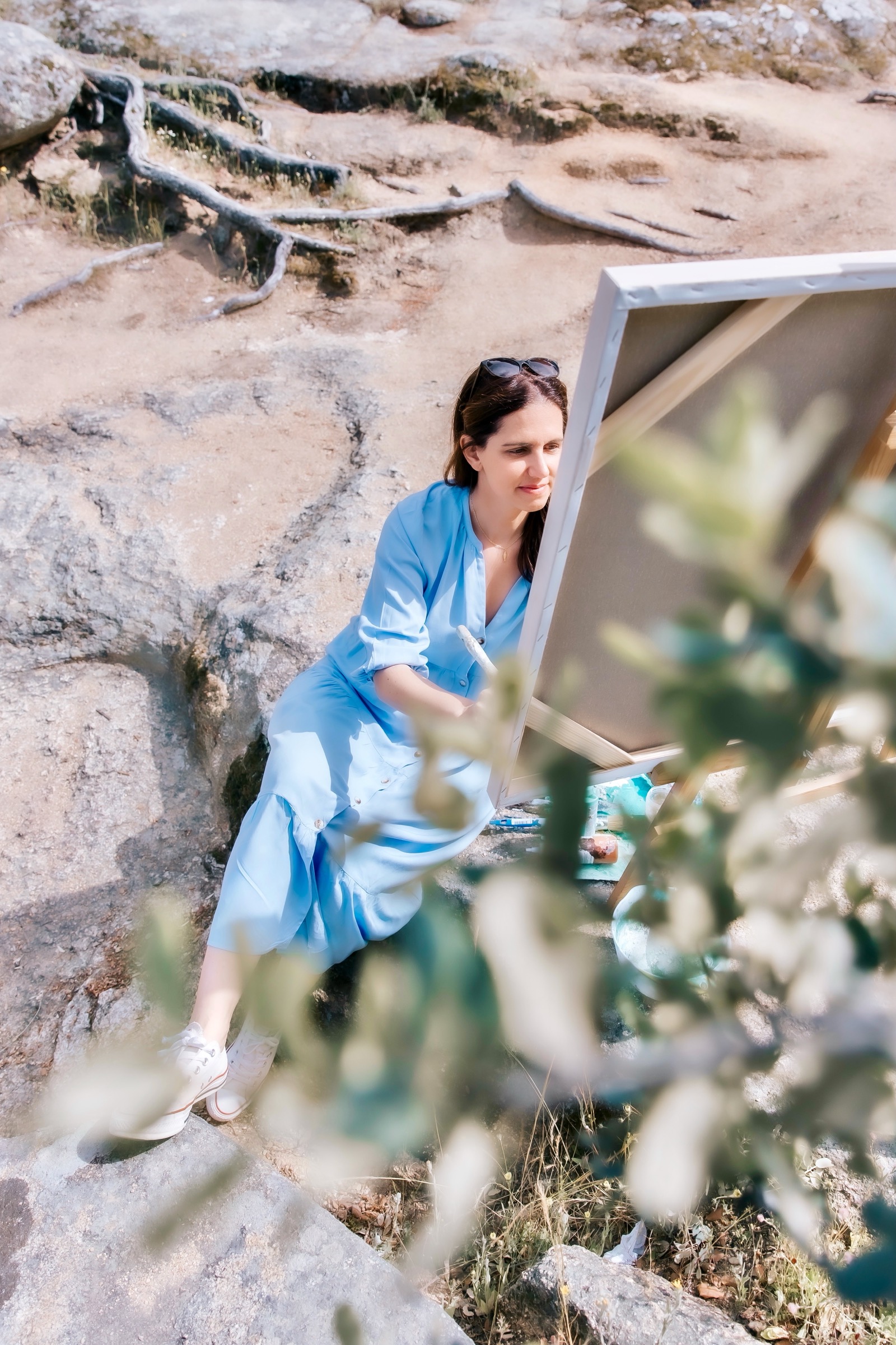 Una persona con vestido azul sentada en rocas al aire libre, trabajando en un lienzo de pintura blanco. La escena está rodeada de naturaleza, con raíces de árboles visibles y vegetación en primer plano.