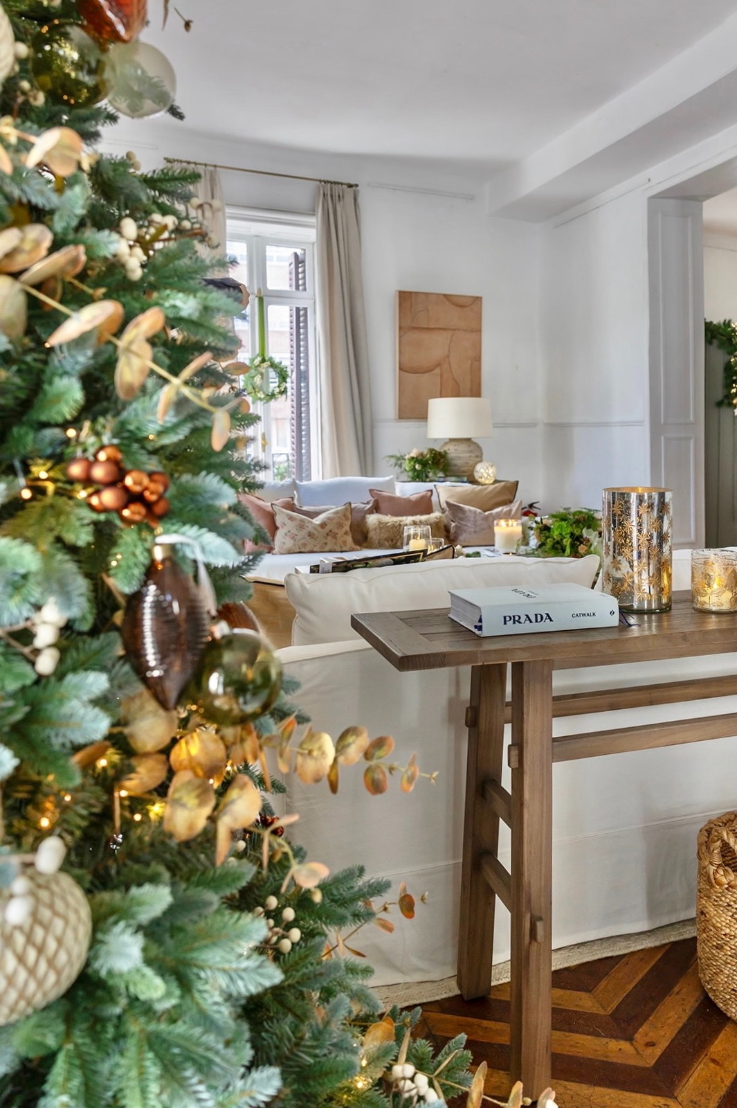 Sala de estar moderna decorada para Navidad con árbol adornado en tonos dorados y cobrizos, mesa consola de madera, sofá blanco con cojines y detalles decorativos elegantes como velas y libros de diseño