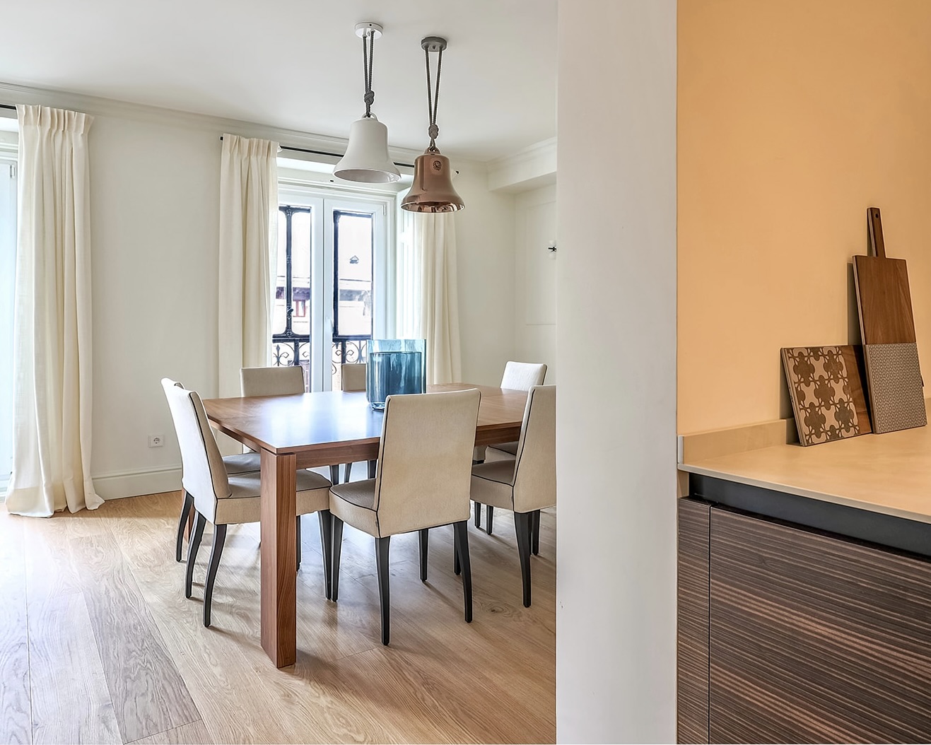 Comedor moderno y elegante con mesa de madera, sillas tapizadas en beige, lámparas colgantes y grandes ventanales con cortinas blancas. El espacio tiene un suelo de madera clara y paredes en tonos neutros.