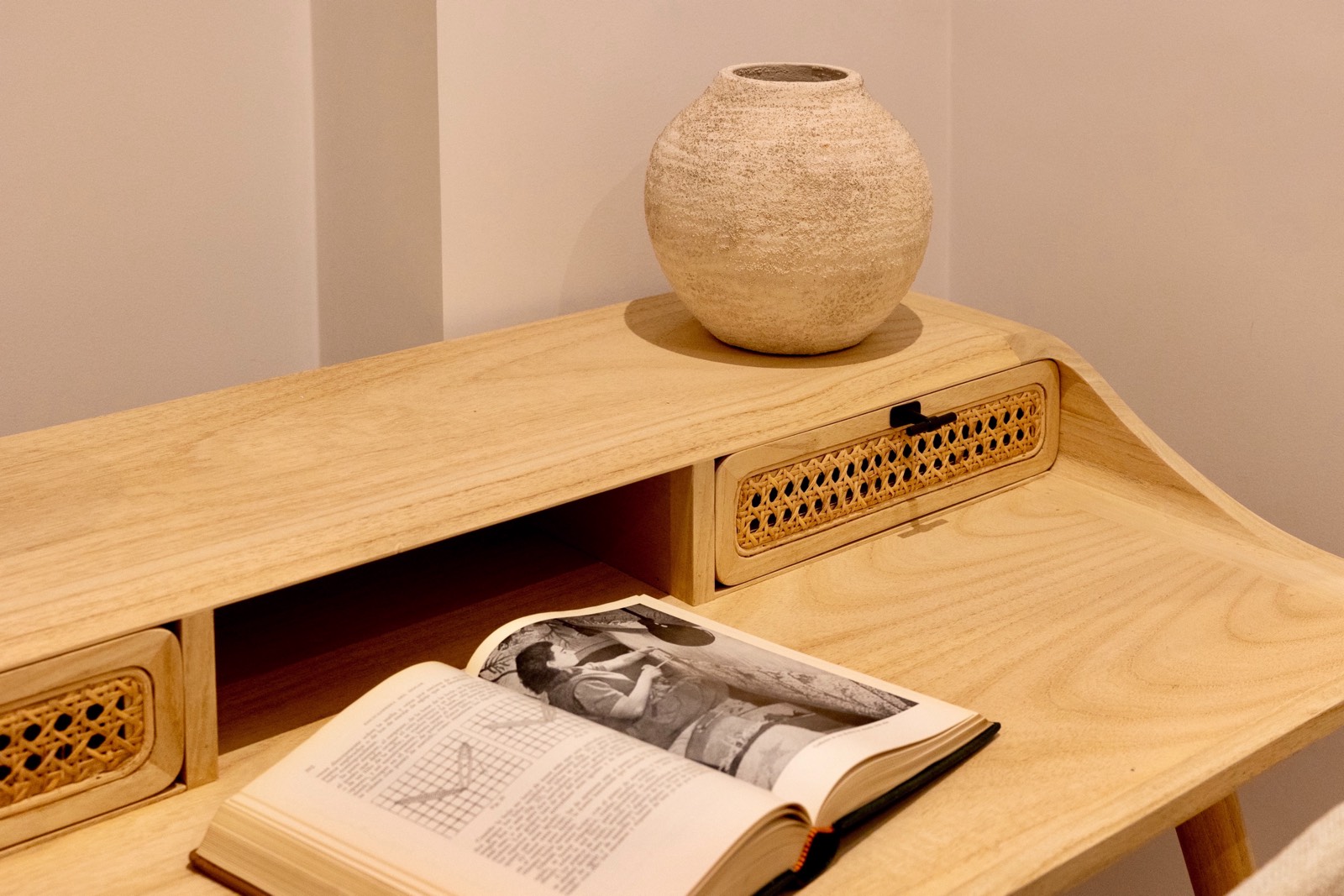 Escritorio de madera con cajones de rejilla, un jarrón esférico de cerámica y un libro abierto mostrando una imagen en blanco y negro. El mueble tiene un diseño moderno y minimalista.