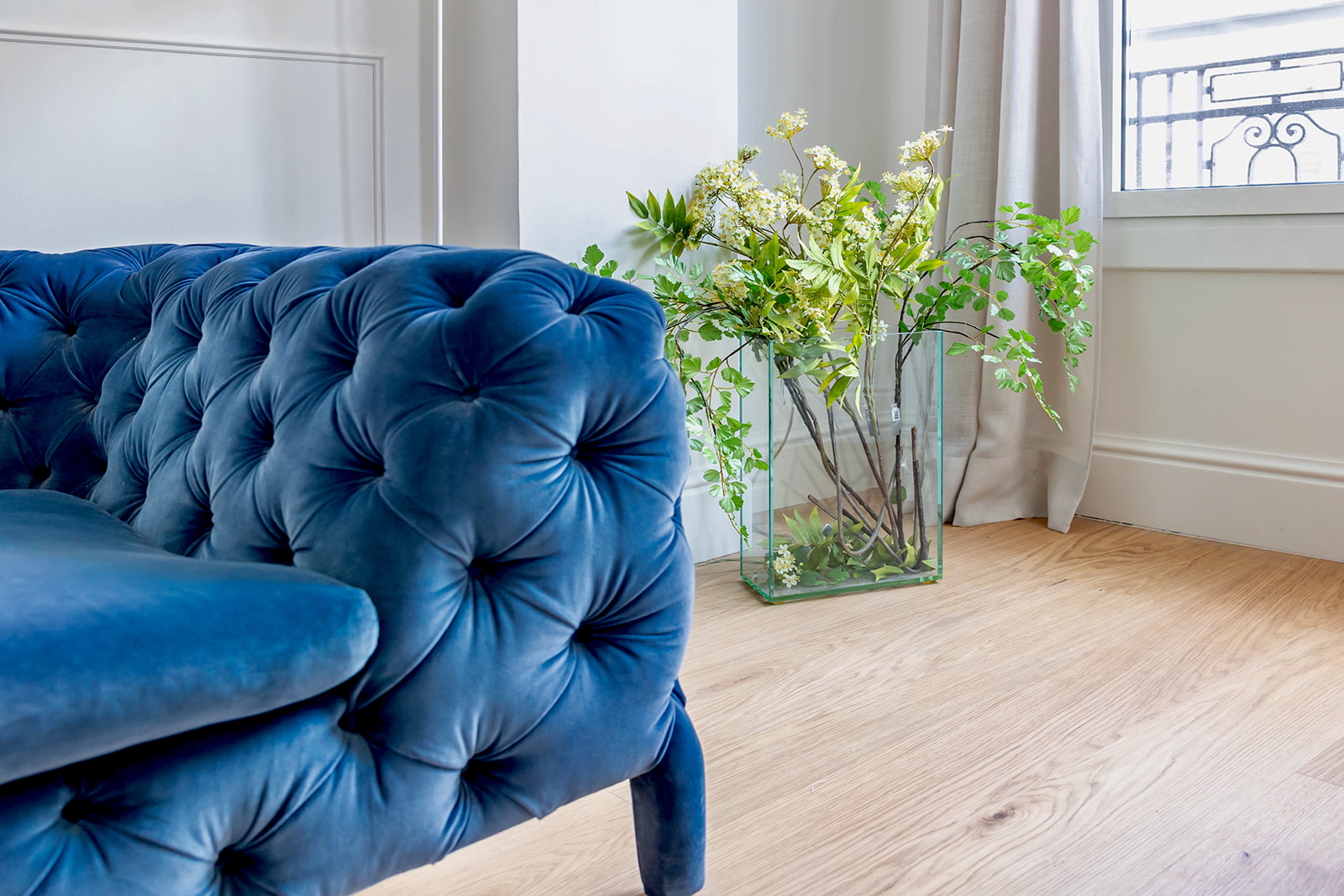 Sofá azul de terciopelo con botones junto a un florero de cristal con flores blancas y hojas verdes en una habitación luminosa con piso de madera y cortinas claras
