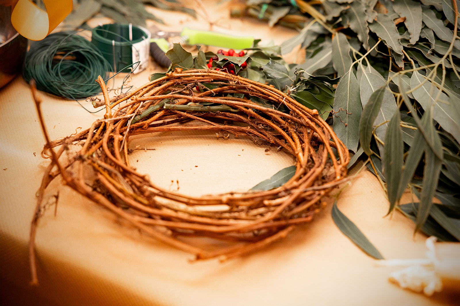 Corona de Navidad hecha de ramas secas rodeada de hojas de eucalipto y materiales para manualidades como hilo verde sobre una superficie de madera