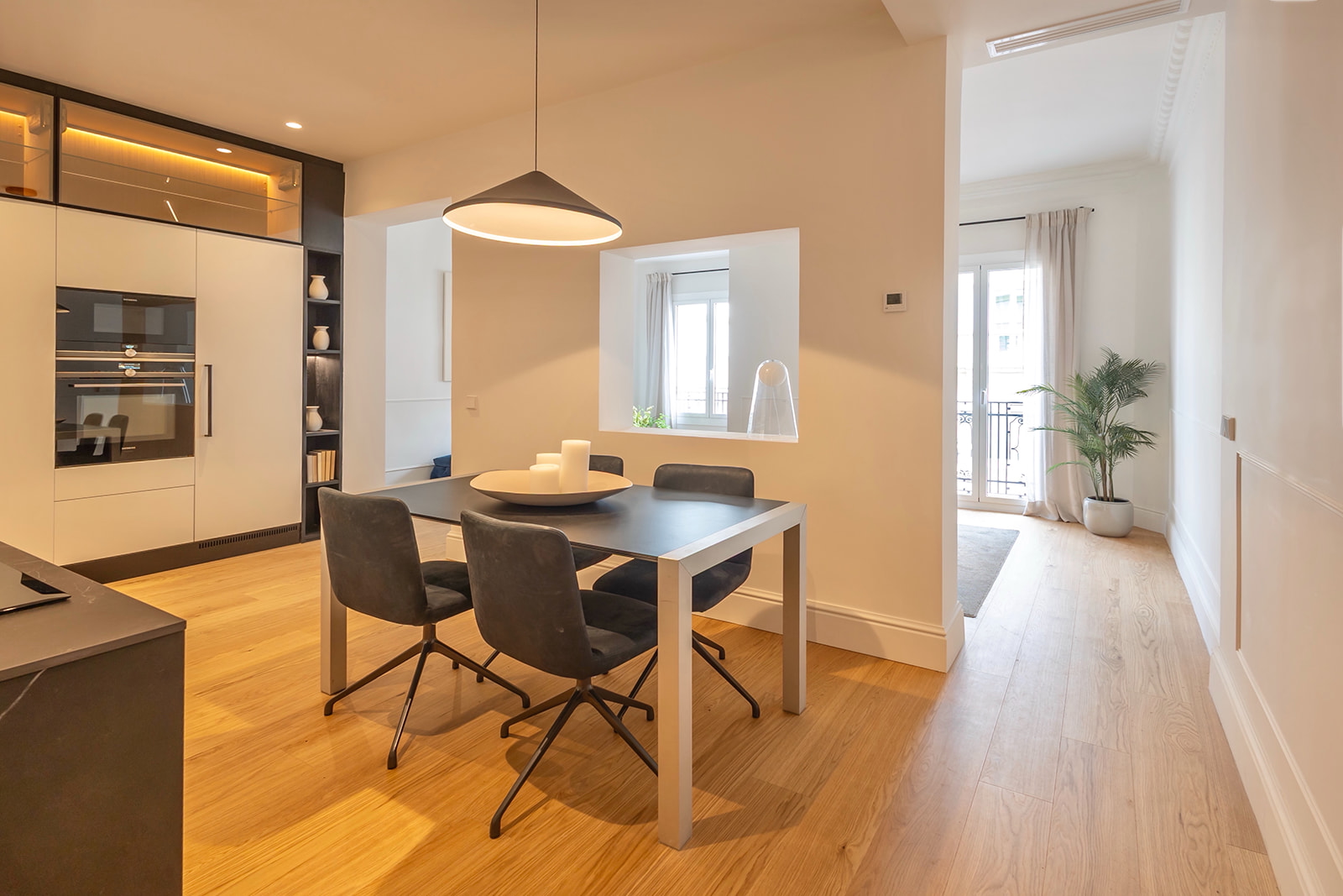 Comedor moderno con mesa rectangular y cuatro sillas negras, integrado en una cocina abierta con electrodomésticos empotrados. La iluminación cálida y el suelo de madera crean un ambiente acogedor.