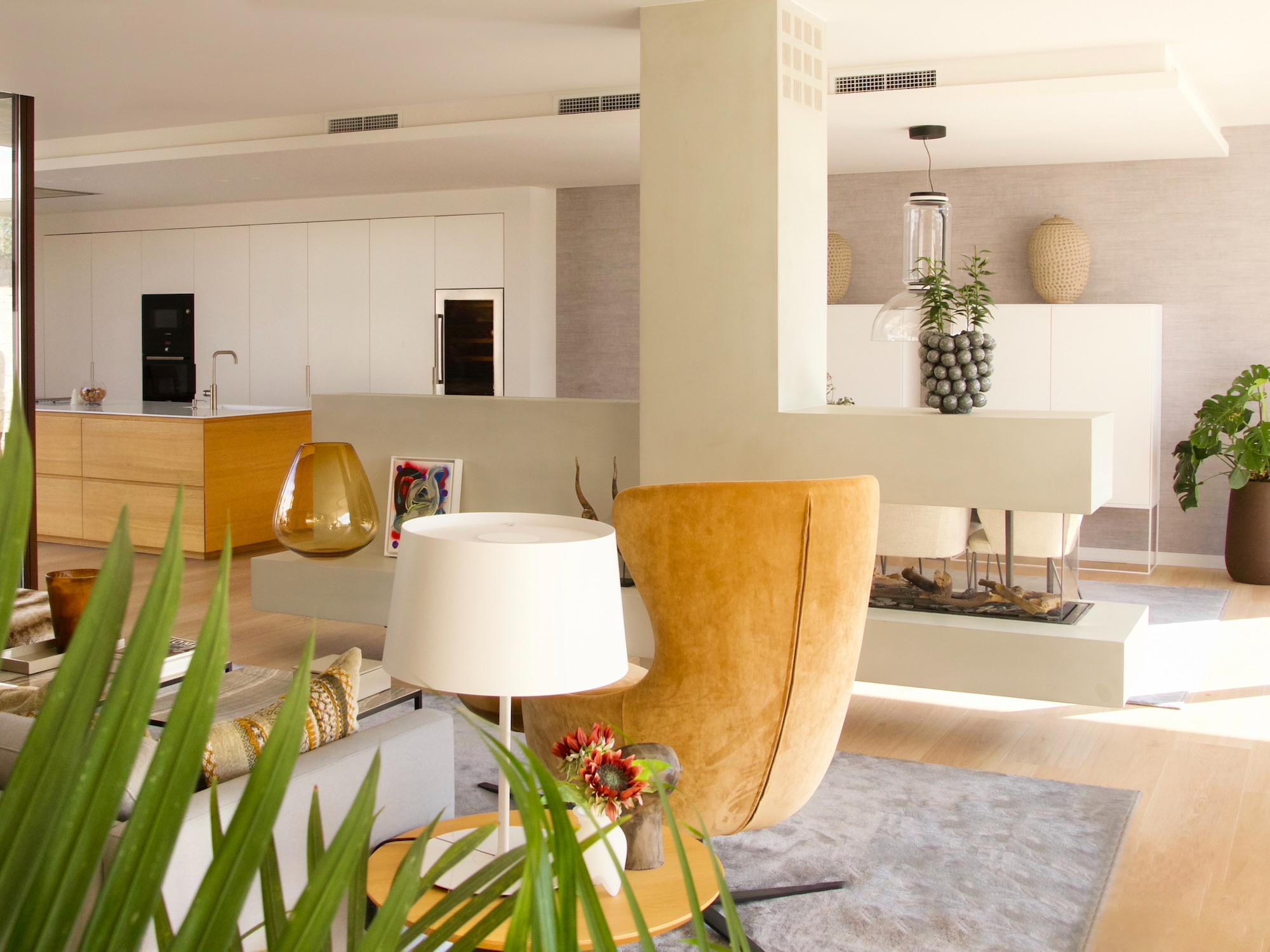 Sala de estar moderna con cocina abierta, decorada en tonos cálidos con sillón mostaza, lámpara blanca, plantas decorativas y mobiliario minimalista. Espacio luminoso con acabados en madera y detalles decorativos contemporáneos.