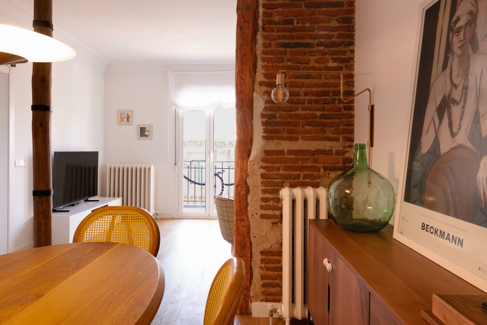 Vista interior de un apartamento con pared de ladrillo, mesa de madera, sillas amarillas, televisor y decoración minimalista