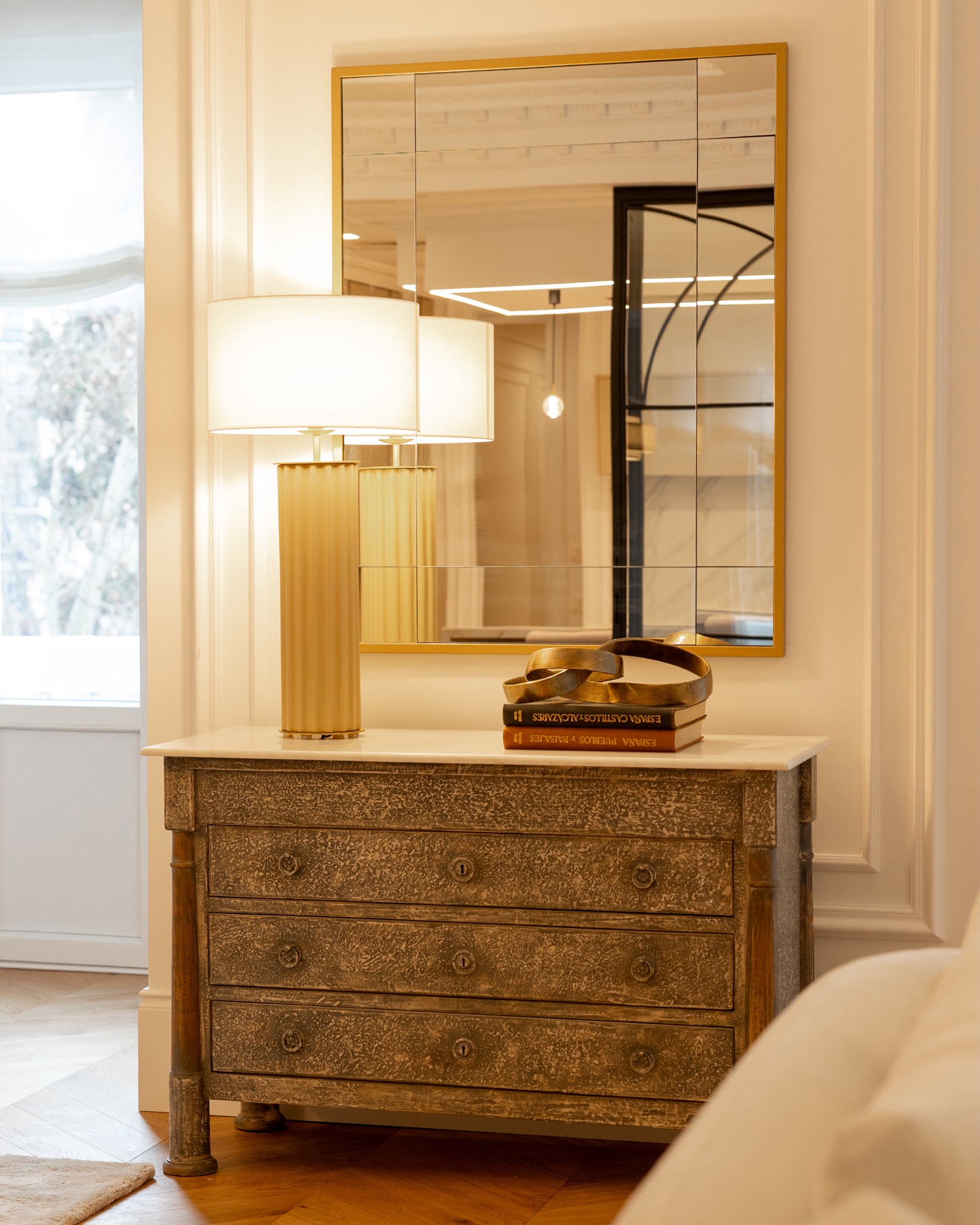 Cómoda antigua de madera con acabado texturizado, espejo cuadrado con marco dorado encima, y lámpara elegante con base dorada y pantalla blanca. Dos libros y un accesorio decorativo sobre la cómoda.