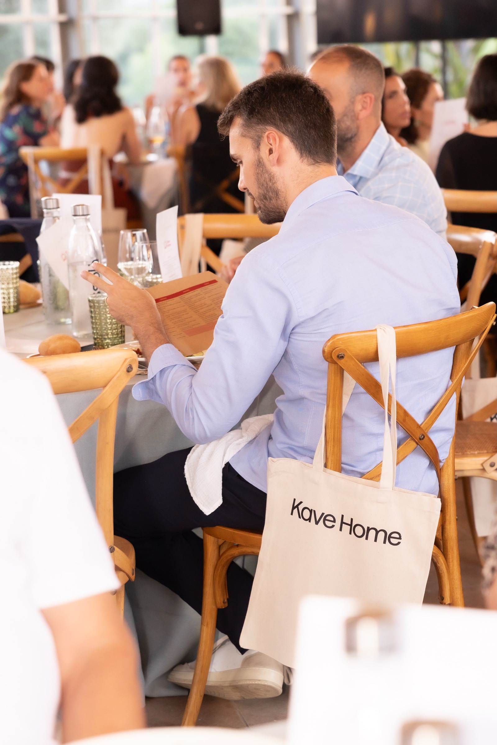 Persona sentada en un restaurante con sillas de madera, sosteniendo un teléfono, con una bolsa de Kave Home en el respaldo de la silla