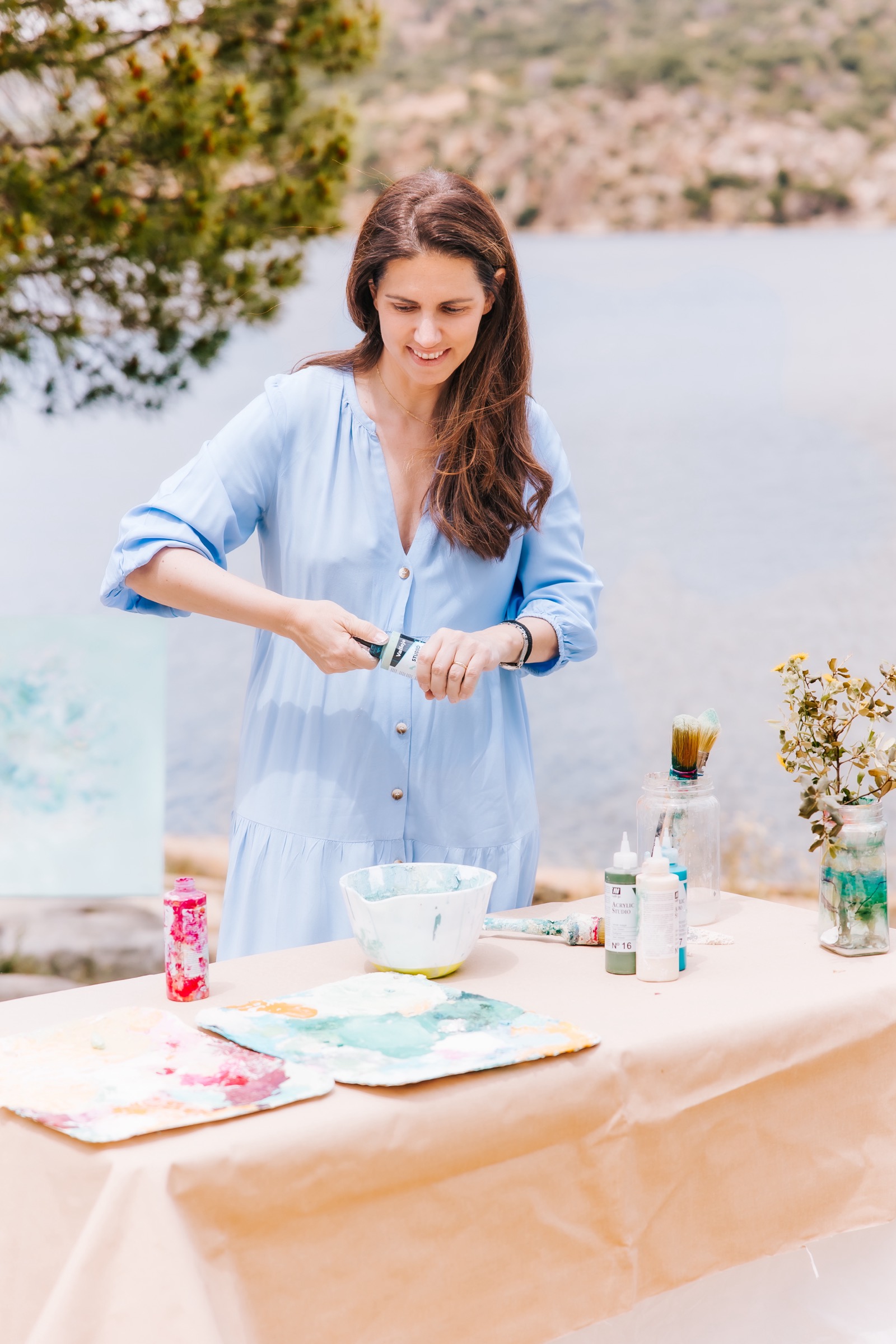 Una mujer en un vestido azul claro pinta al aire libre junto a un lago. Sobre una mesa hay materiales de arte como pinturas y pinceles. El fondo muestra un hermoso paisaje de montaña y agua.