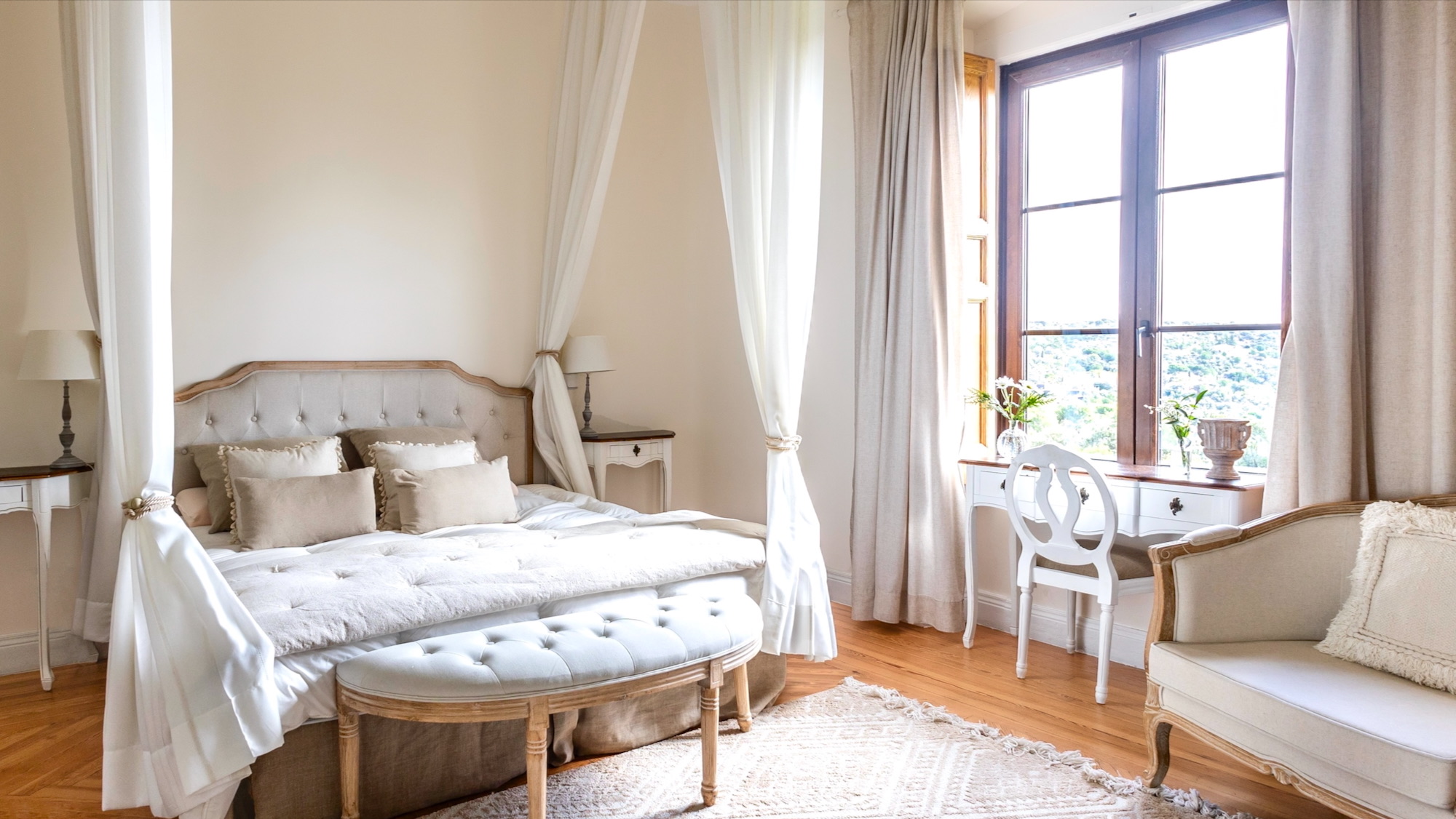 Dormitorio elegante en tonos blancos y beige con cama dosel, banco tapizado al pie de la cama, escritorio junto a ventana con vistas y mobiliario de estilo provenzal sobre suelo de parquet
