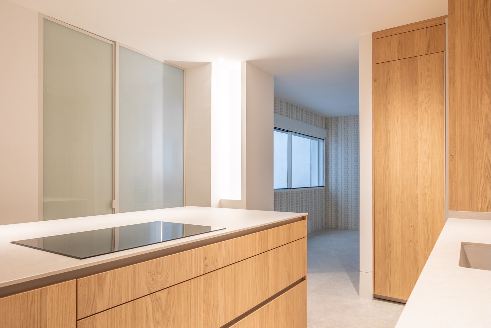 Interior de cocina minimalista con isla central, muebles de madera clara, encimera blanca y grandes ventanales