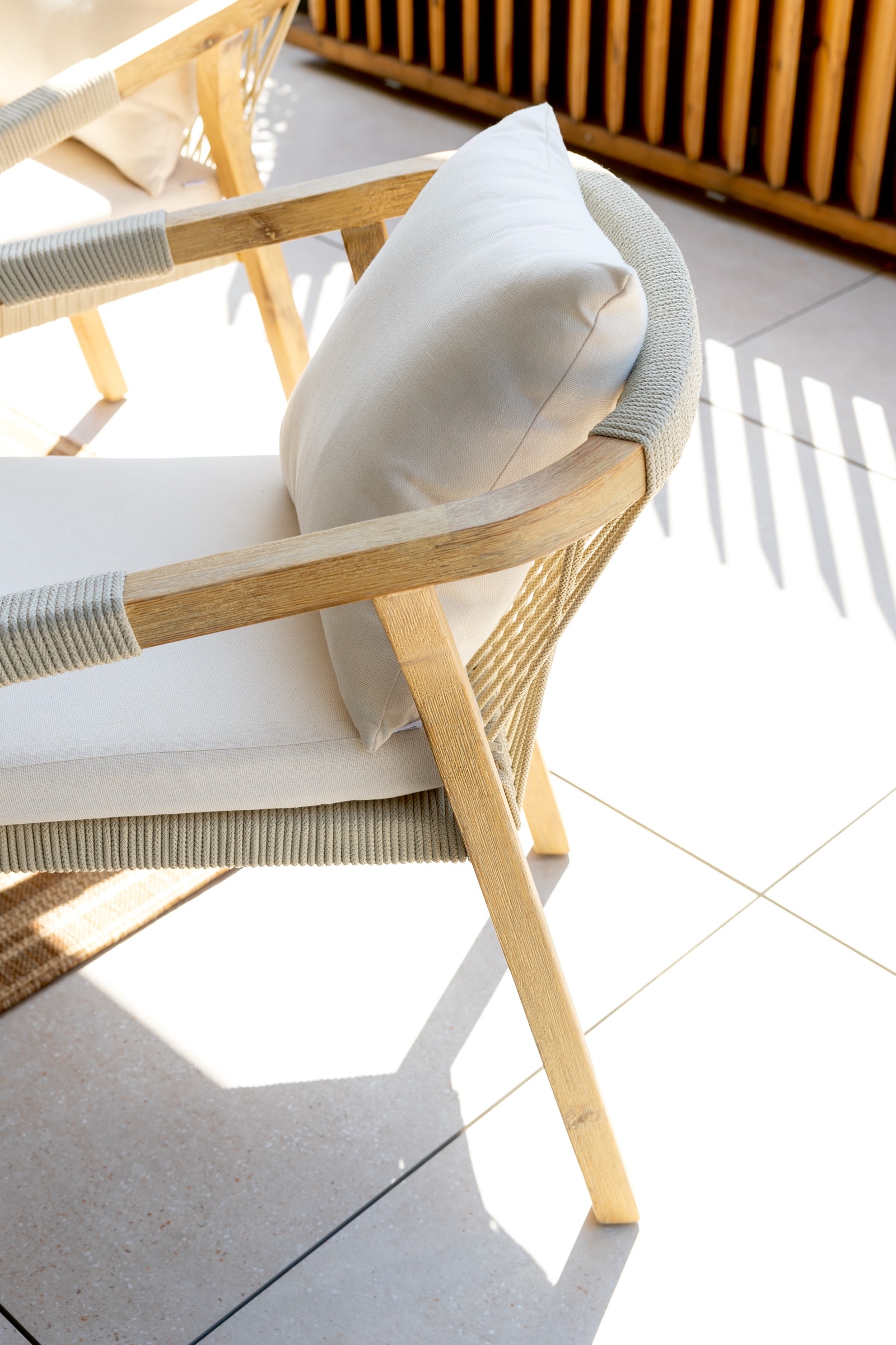 Silla de diseño escandinavo con estructura de madera clara y cojín blanco, ubicada en una terraza soleada con suelo de baldosas. La silla muestra detalles de cuerda trenzada y está iluminada por la luz natural.