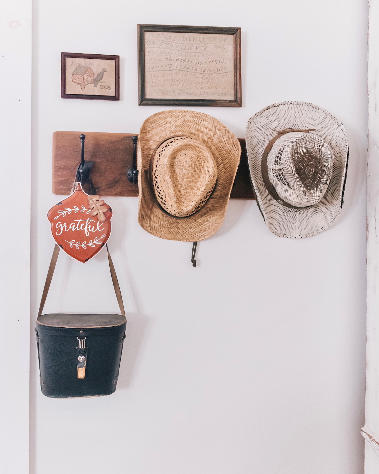 Perchero de madera en pared blanca con dos sombreros vaqueros, uno de paja y otro gris, un bolso negro colgado y un adorno decorativo que dice 'grateful'. Arriba hay dos cuadros enmarcados en la pared.