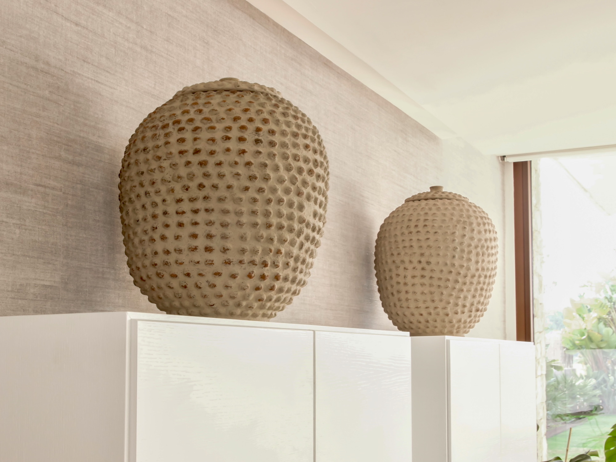 Dos jarrones decorativos grandes de cerámica en color beige con textura de puntos en relieve, colocados sobre muebles blancos contra una pared clara en un ambiente minimalista y moderno