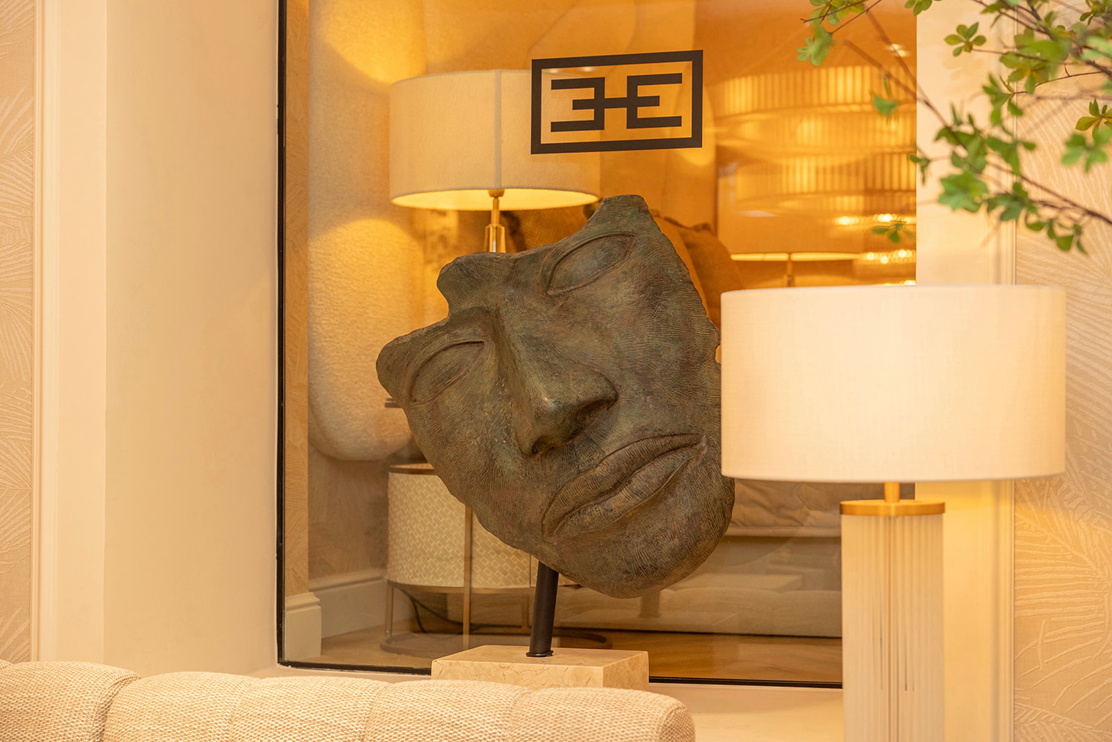 Escultura de bronce de un rostro abstracto en un ambiente interior elegante con lámparas y decoración en tonos cálidos. Logo 'EE' visible en el fondo.