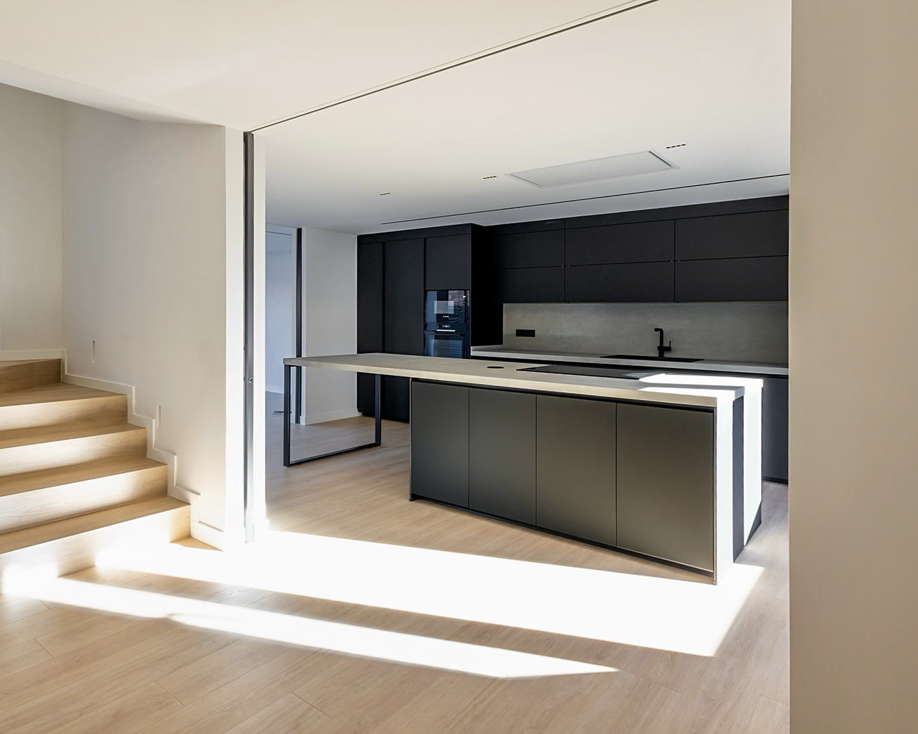 Cocina moderna y minimalista con isla central iluminada, gabinetes negros, electrodomésticos integrados y piso de madera clara. Escaleras adyacentes y diseño abierto con mucha luz natural.