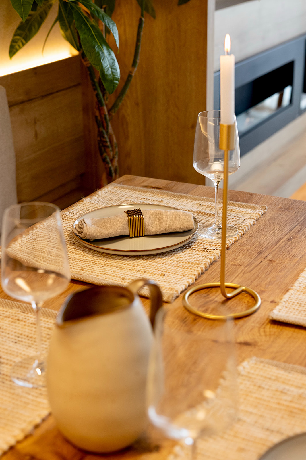 Mesa de madera con mantel individual de yute, plato con servilleta, copas de vino, candelabro dorado con vela blanca encendida y jarra blanca. Ambiente cálido y acogedor con planta en el fondo.