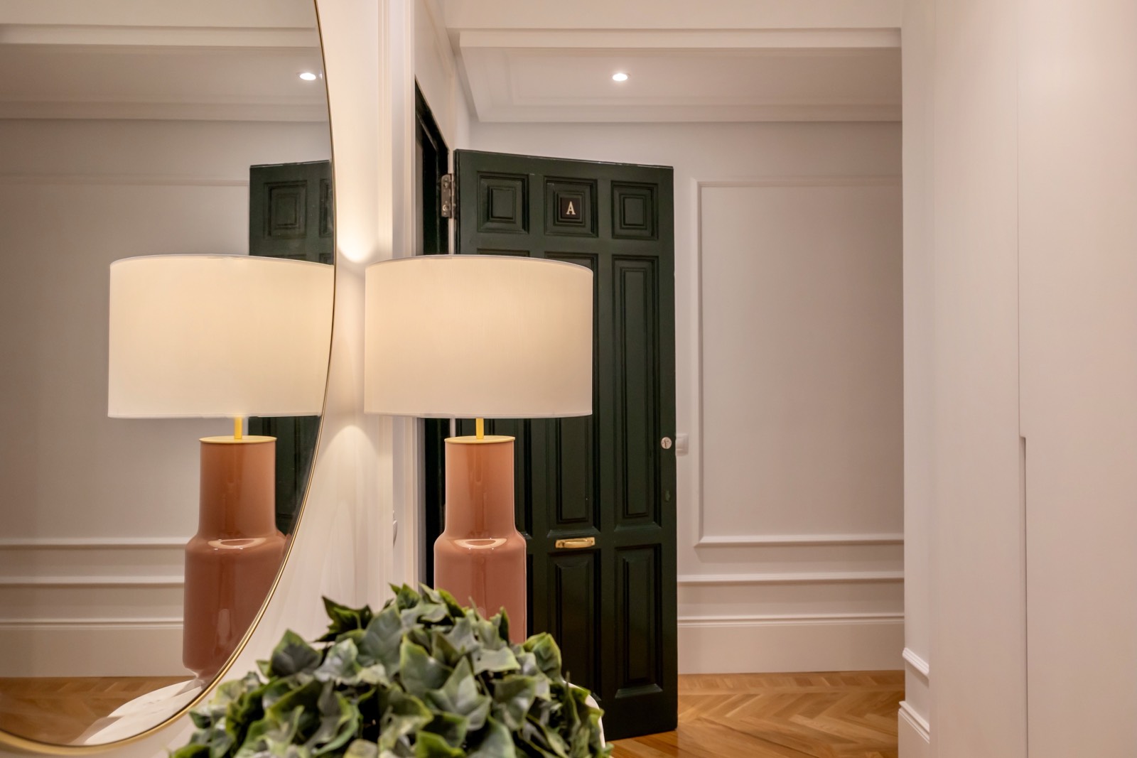 Elegante entrada de una casa con una lámpara de mesa rosa y pantalla blanca, reflejada en un espejo. Al fondo se ve una puerta verde oscuro con paneles. El espacio tiene paredes blancas y suelo de parquet.