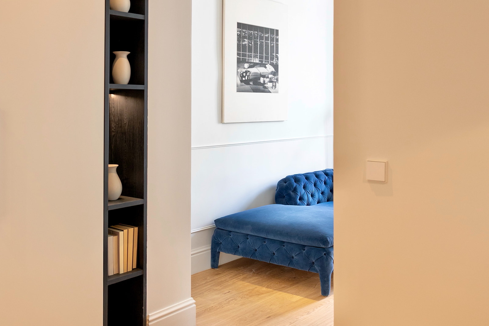 Vista parcial de una habitación elegante con un sofá azul capitoné, una estantería oscura con jarrones y libros, y una imagen enmarcada en la pared blanca. El suelo es de madera clara.