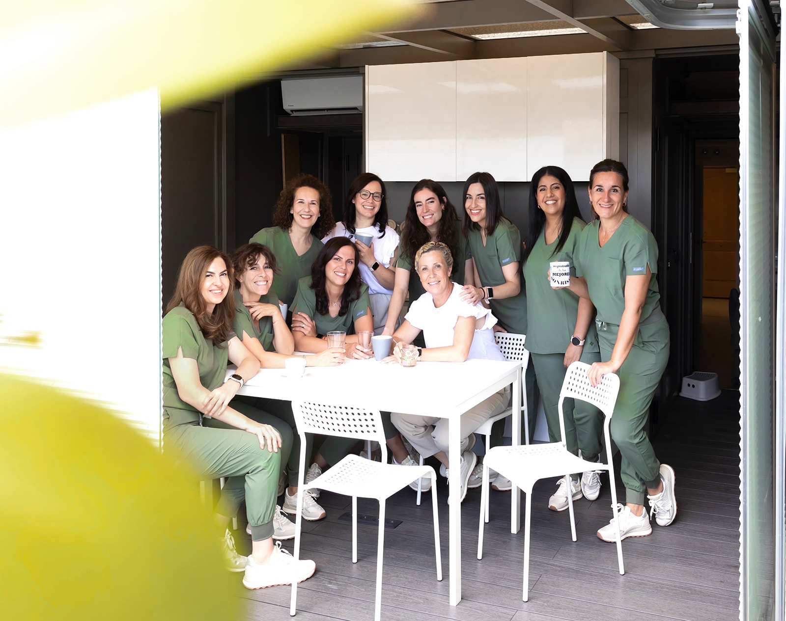 Grupo de profesionales médicas en uniformes verdes reunidas alrededor de una mesa blanca en un área de descanso, sonriendo y compartiendo un momento de camaradería