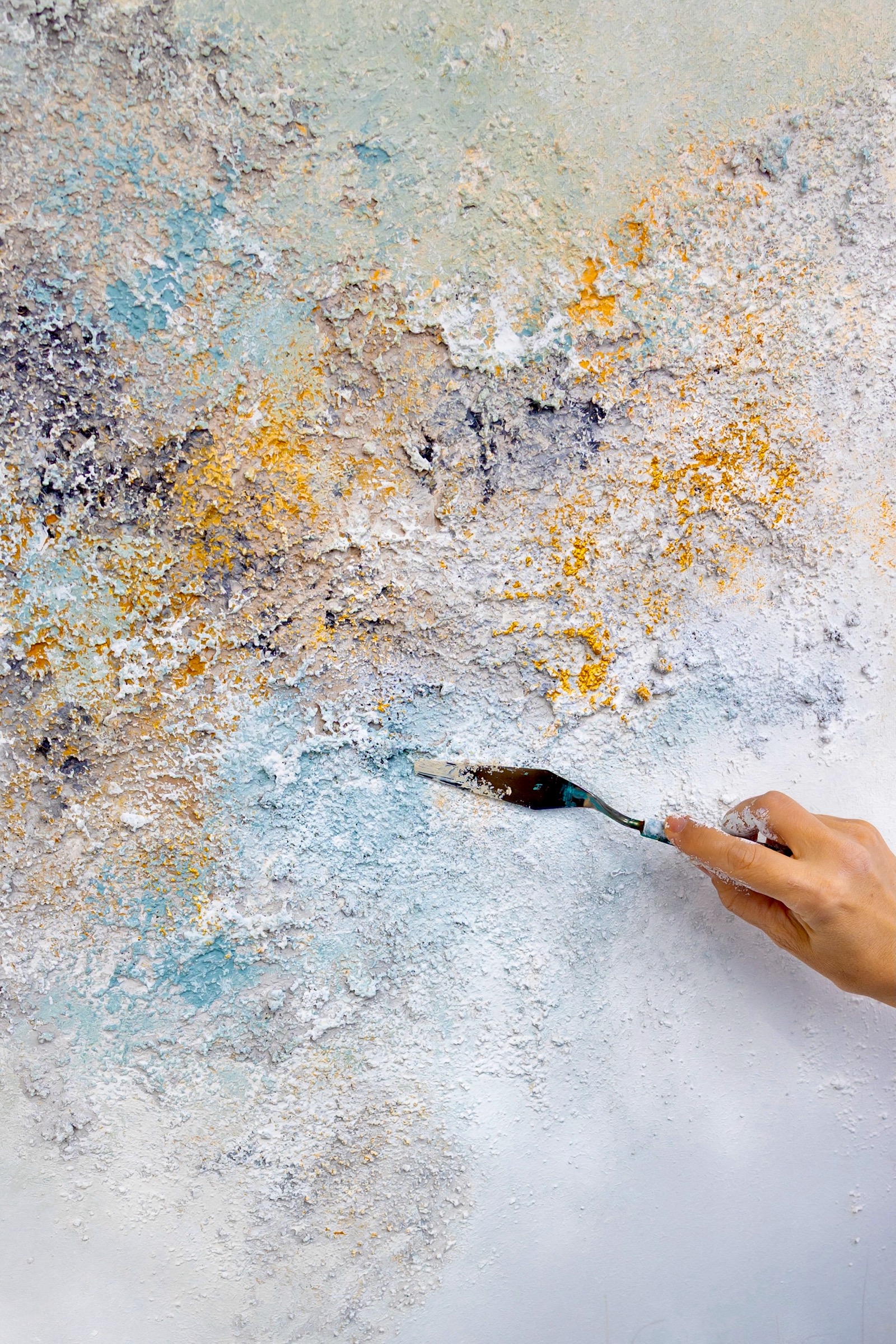 Mano sosteniendo un cuchillo de pintura aplicando textura a una superficie abstracta con colores pastel azul, amarillo y blanco
