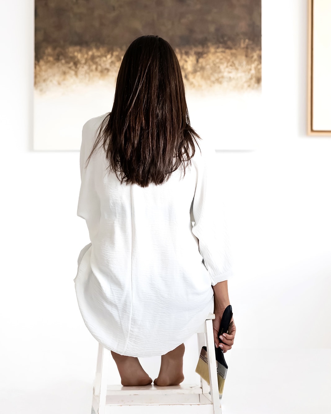 Vista trasera de una mujer con cabello largo oscuro, vistiendo una camisa blanca suelta, sentada en una escalera blanca mientras observa una obra de arte abstracta en una galería