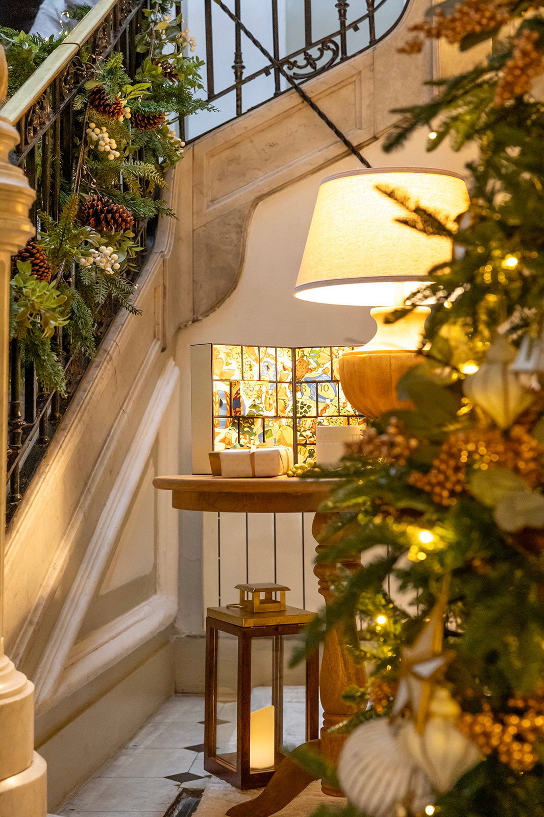 Decoración navideña en escalera interior con guirnaldas naturales, piñas, lámpara de mesa con pantalla blanca, farol de suelo y detalles dorados iluminados creando una atmósfera cálida y acogedora