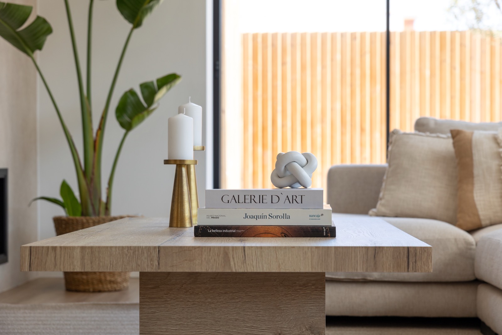 Mesa de centro de madera con libros de arte, velas y una escultura abstracta. Al fondo se ve un sofá beige y una planta grande en una maceta de mimbre. La habitación tiene un estilo moderno y minimalista.