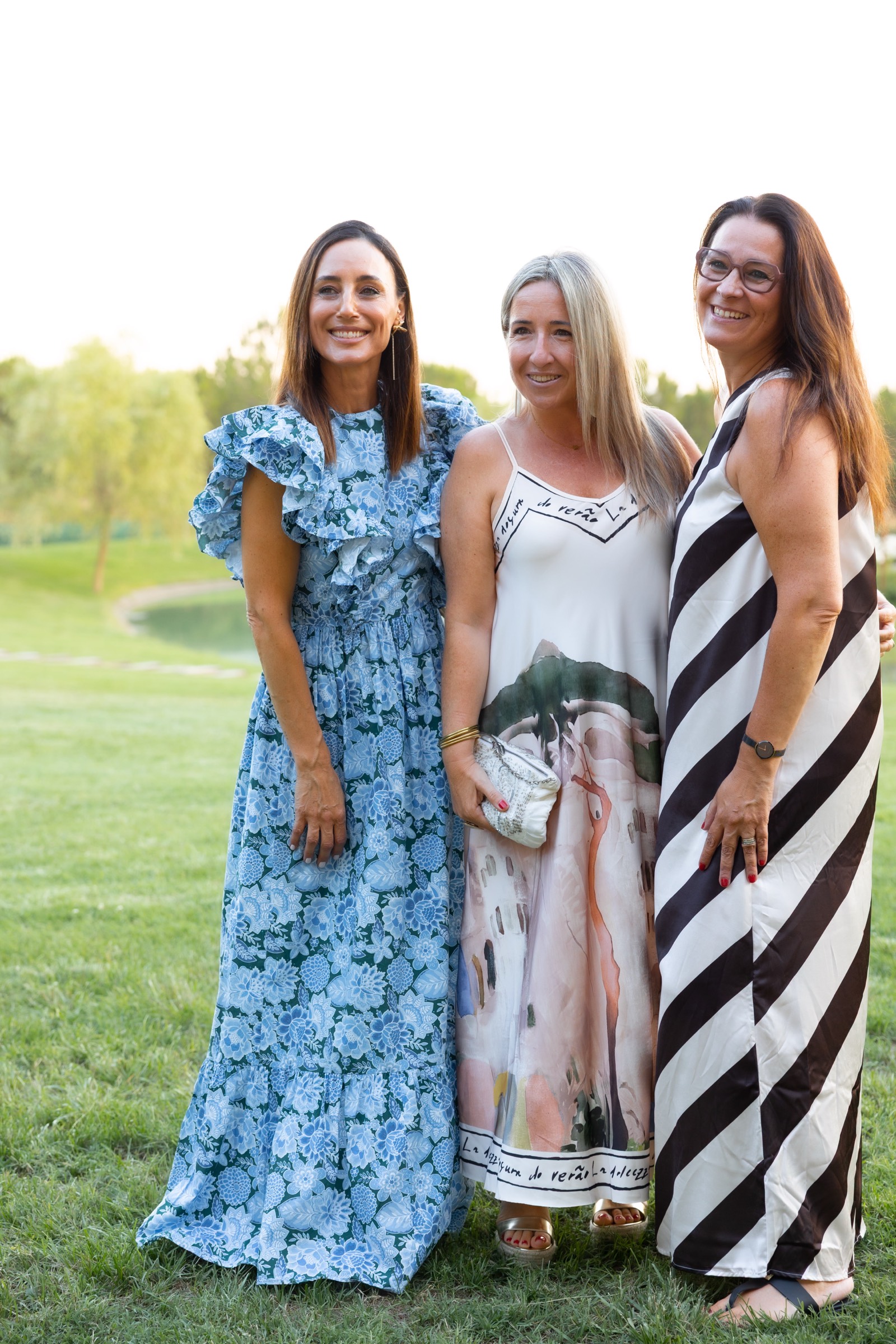 Tres mujeres sonrientes de pie en un jardín verde, usando vestidos largos elegantes en azul, blanco y blanco y negro