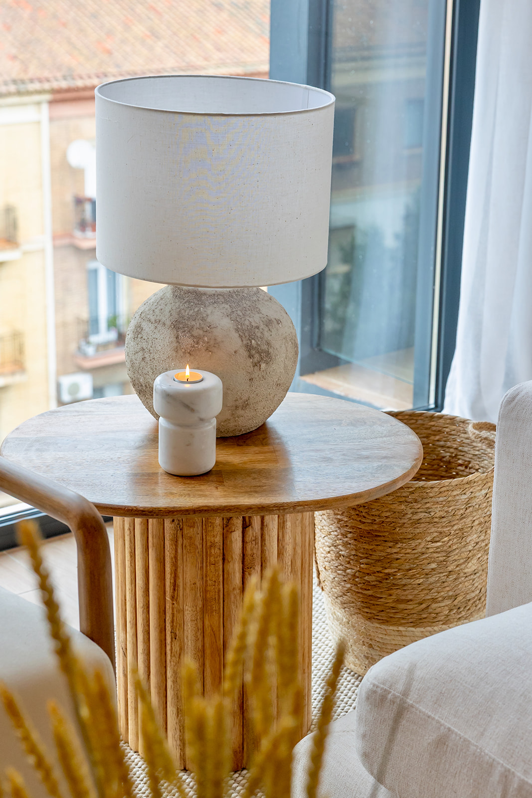 Lámpara de mesa con base redonda de piedra y pantalla blanca sobre una mesa auxiliar de madera redonda. Una vela encendida en un portavelas de mármol blanco y una cesta de fibra natural completan la escena junto a una ventana.