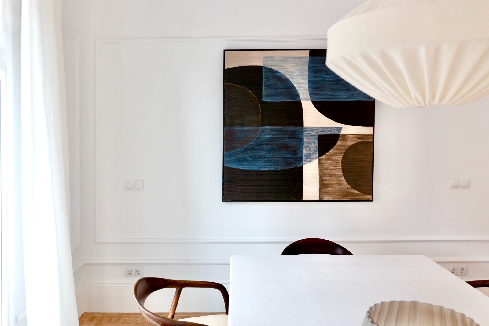 Comedor moderno con paredes blancas, mesa blanca y sillas de madera. En la pared, un cuadro abstracto grande con formas geométricas en tonos azules, negros y marrones. Una lámpara de techo blanca y redonda ilumina la escena.
