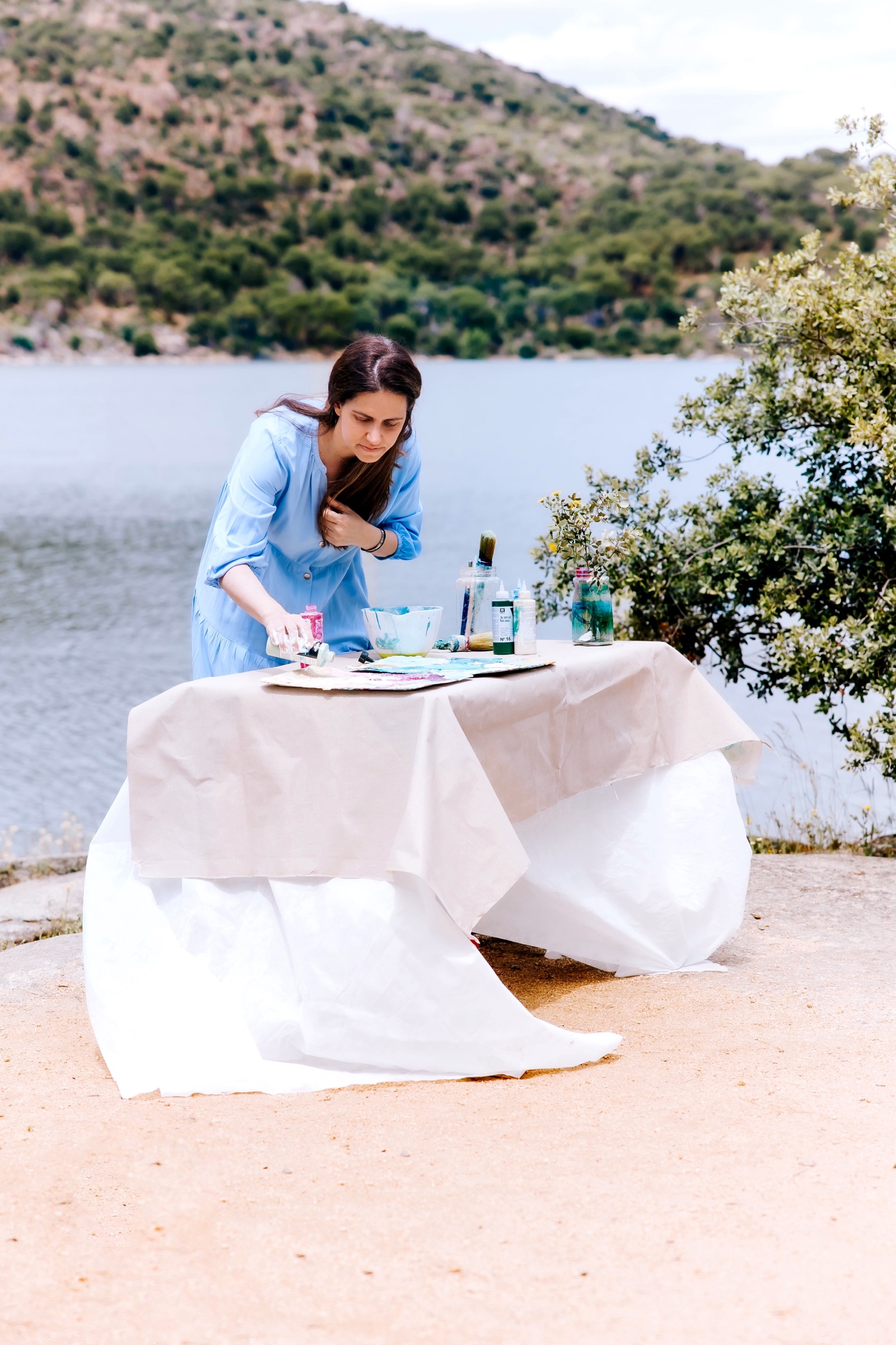 Escena de picnic junto a un lago tranquilo. Una mesa cubierta con mantel blanco está puesta con platos y bebidas. El fondo muestra colinas y vegetación alrededor del agua.
