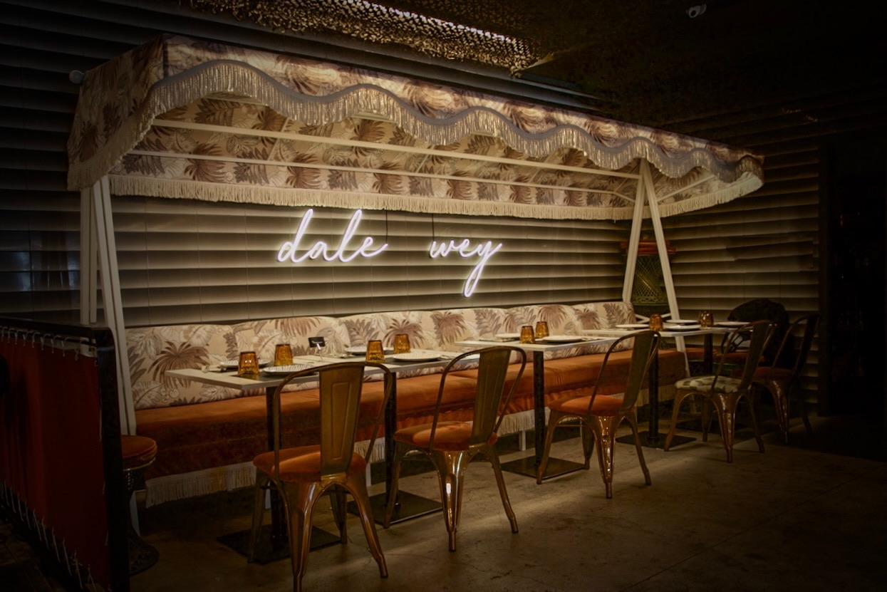 Interior de restaurante con letrero de neón 'dale wey', decoración tropical con estampados de palmeras, asientos tipo cabina tapizados en naranja, sillas metálicas y ambiente acogedor con iluminación tenue