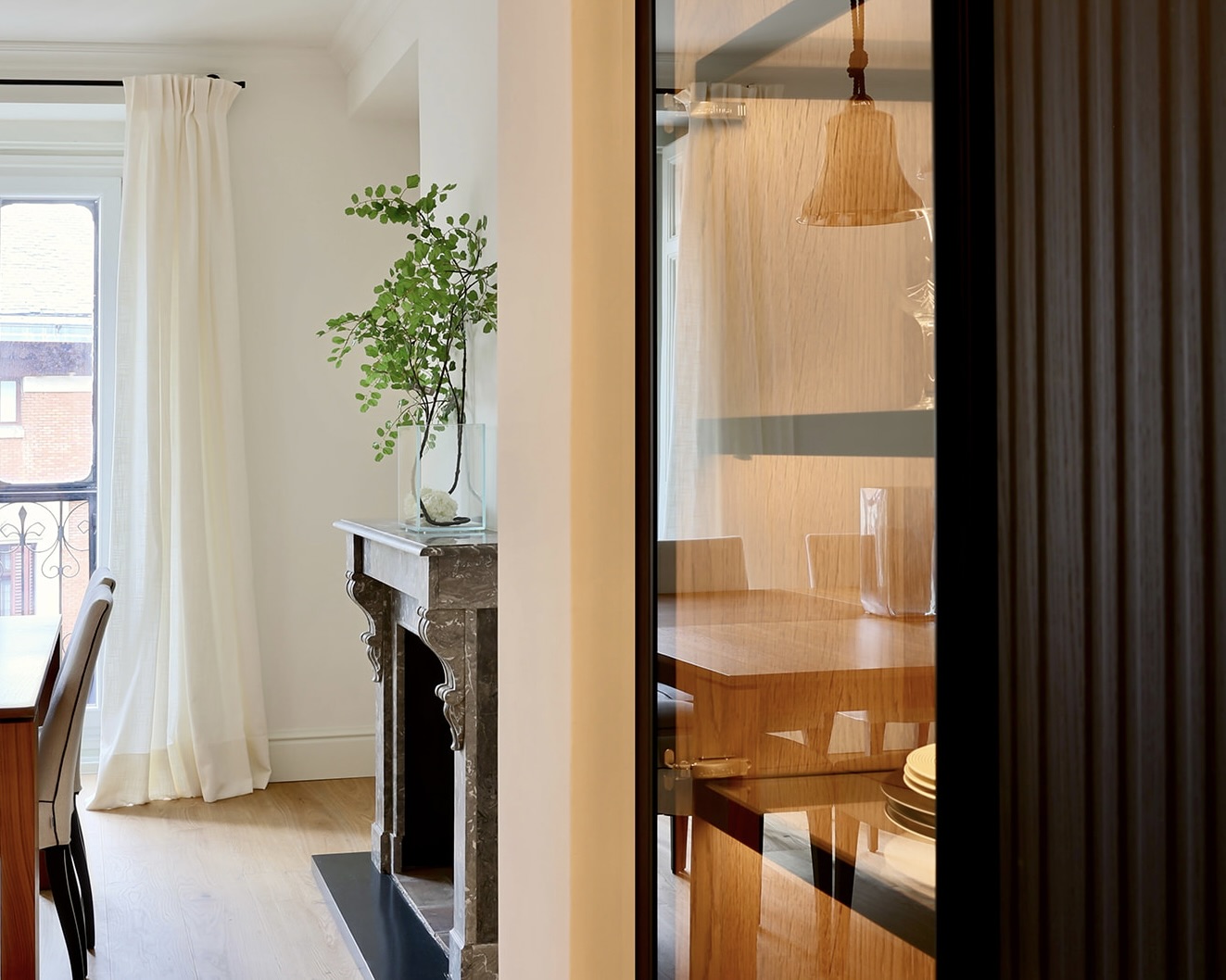 Interior elegante con chimenea de mármol y planta verde. Vista parcial de comedor con mesa de madera y lámpara colgante. Ventana con cortinas blancas y suelo de madera.