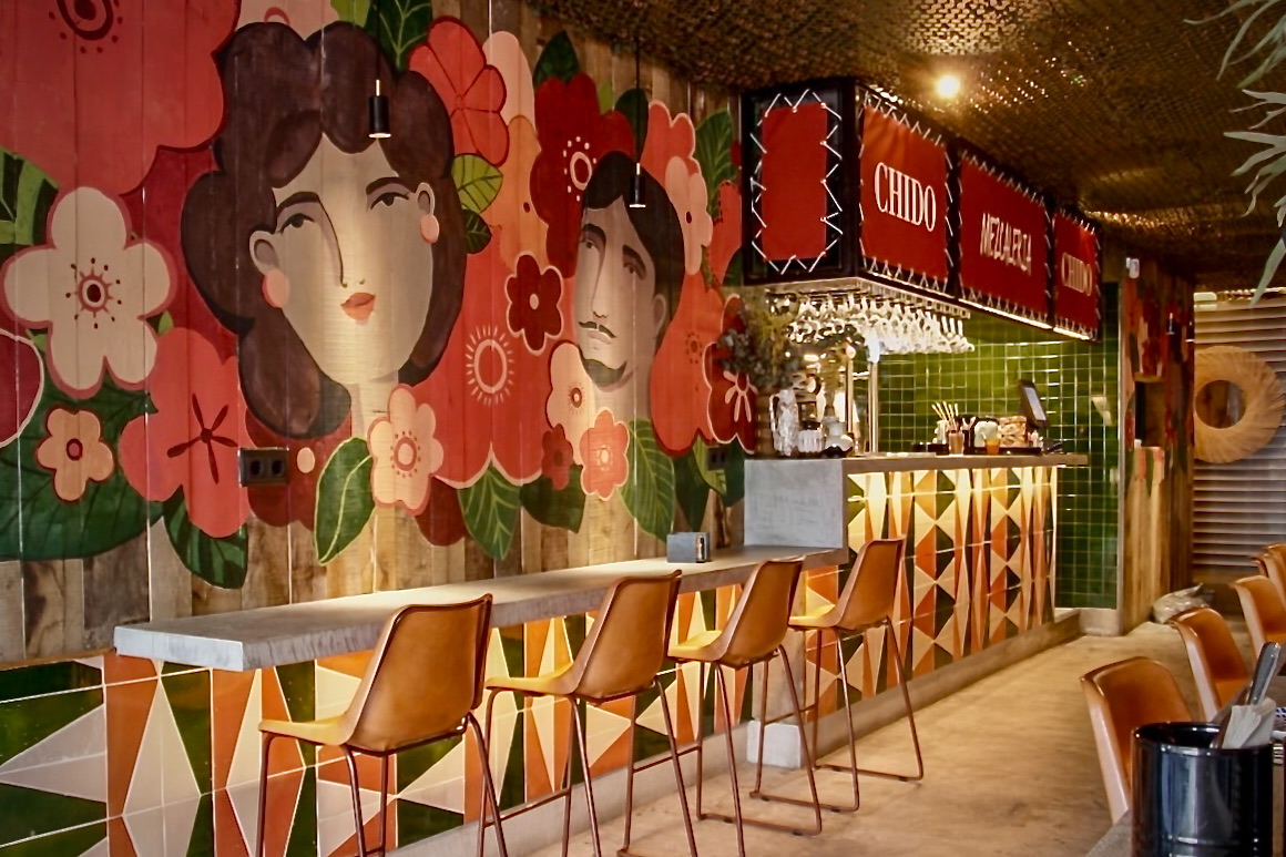 Interior de restaurante mexicano con mural artístico floral en tonos coral y rosa, barra con sillas altas color caramelo y decoración geométrica en la base. Iluminación cálida y ambiente acogedor.