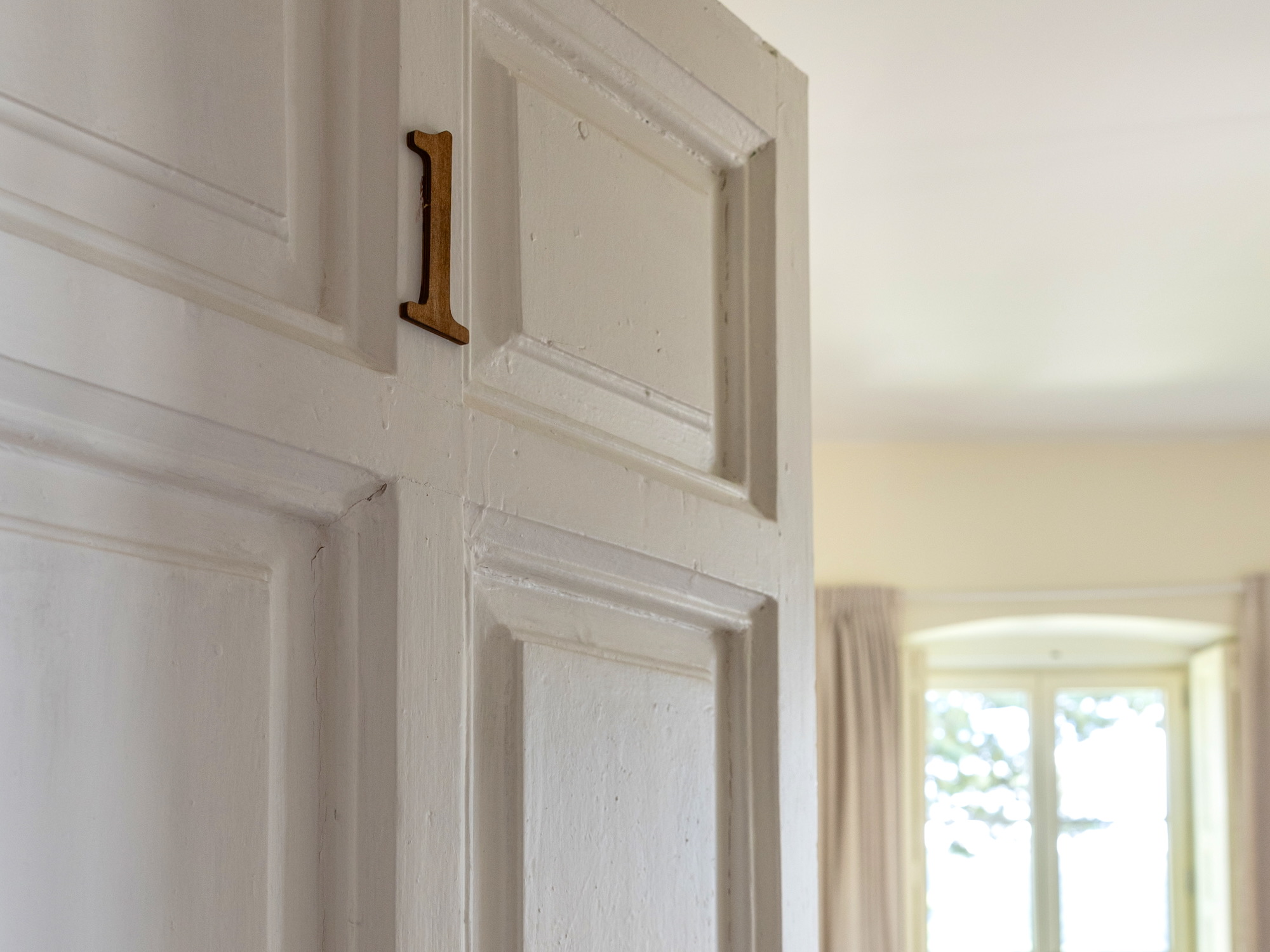 Primer plano de una puerta blanca con un número 1 de madera montado sobre ella. Al fondo se aprecia una habitación luminosa con cortinas y una ventana.