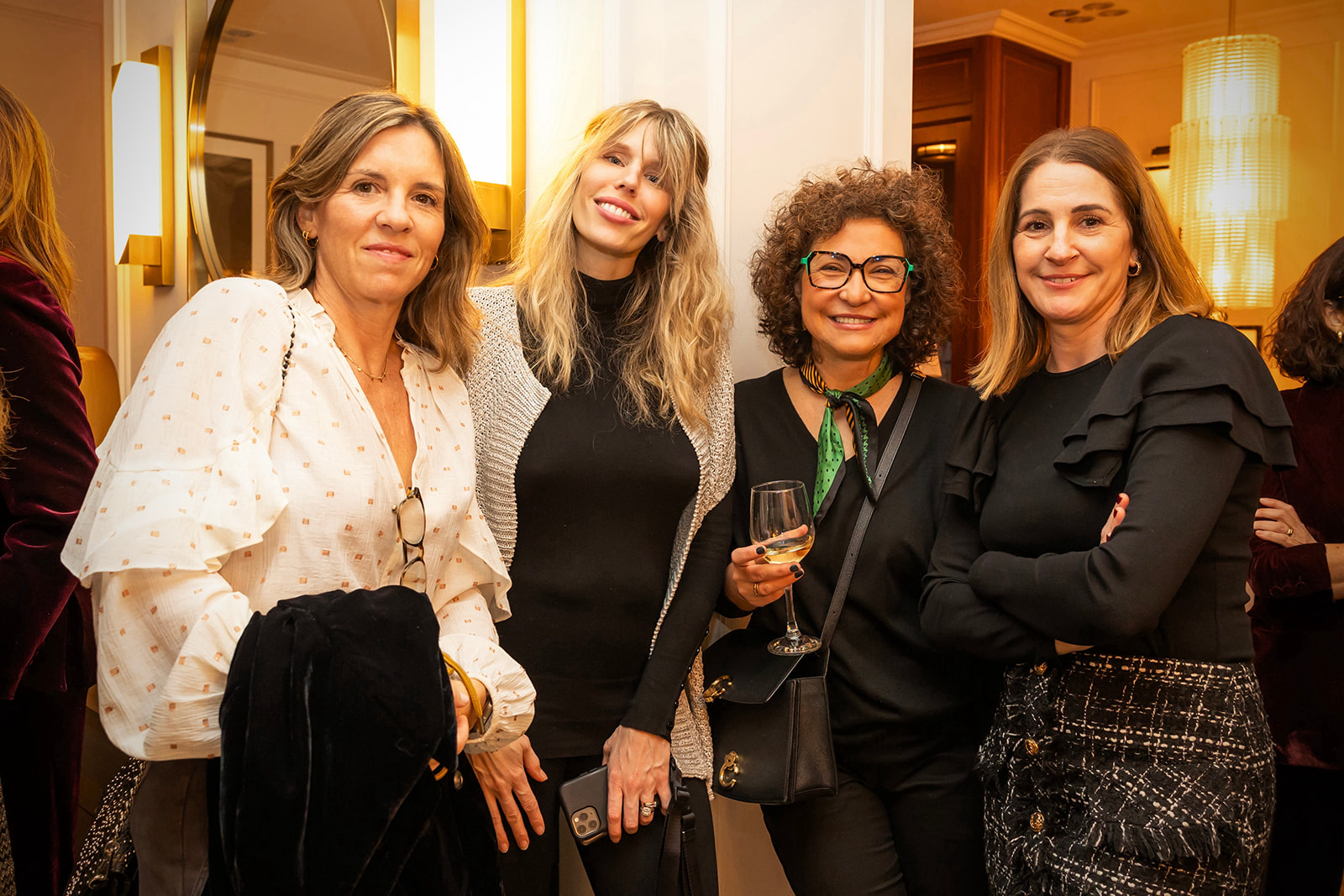 Cuatro mujeres sonrientes posando juntas en un evento social elegante. El ambiente es cálido y acogedor, con iluminación suave y decoración lujosa. Las mujeres visten ropa elegante y sostienen bebidas.
