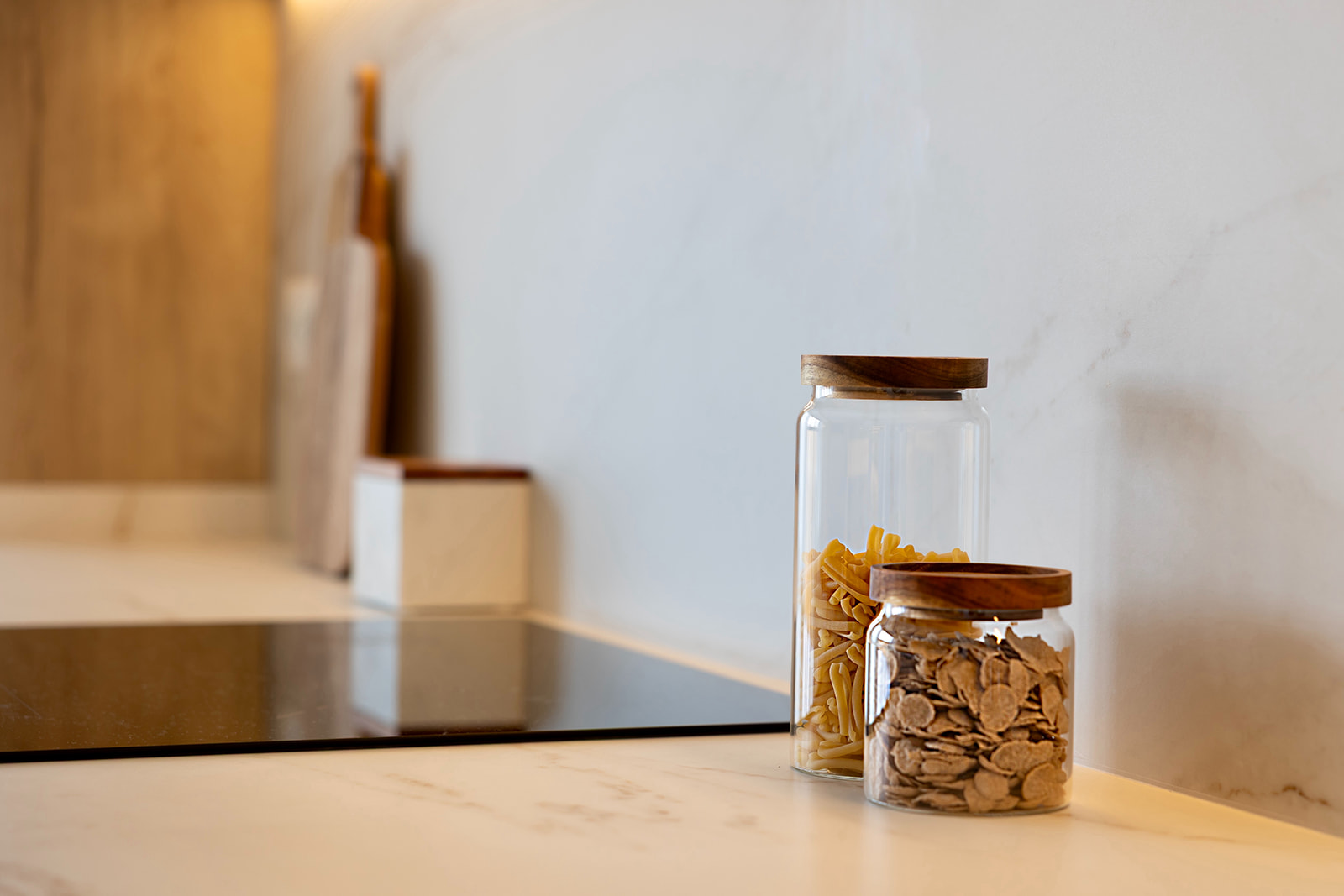 Dos frascos de vidrio con tapas de madera en una encimera de cocina. Uno contiene pasta y el otro cereales. Al fondo se ve una superficie de cocina y otros elementos de almacenamiento.