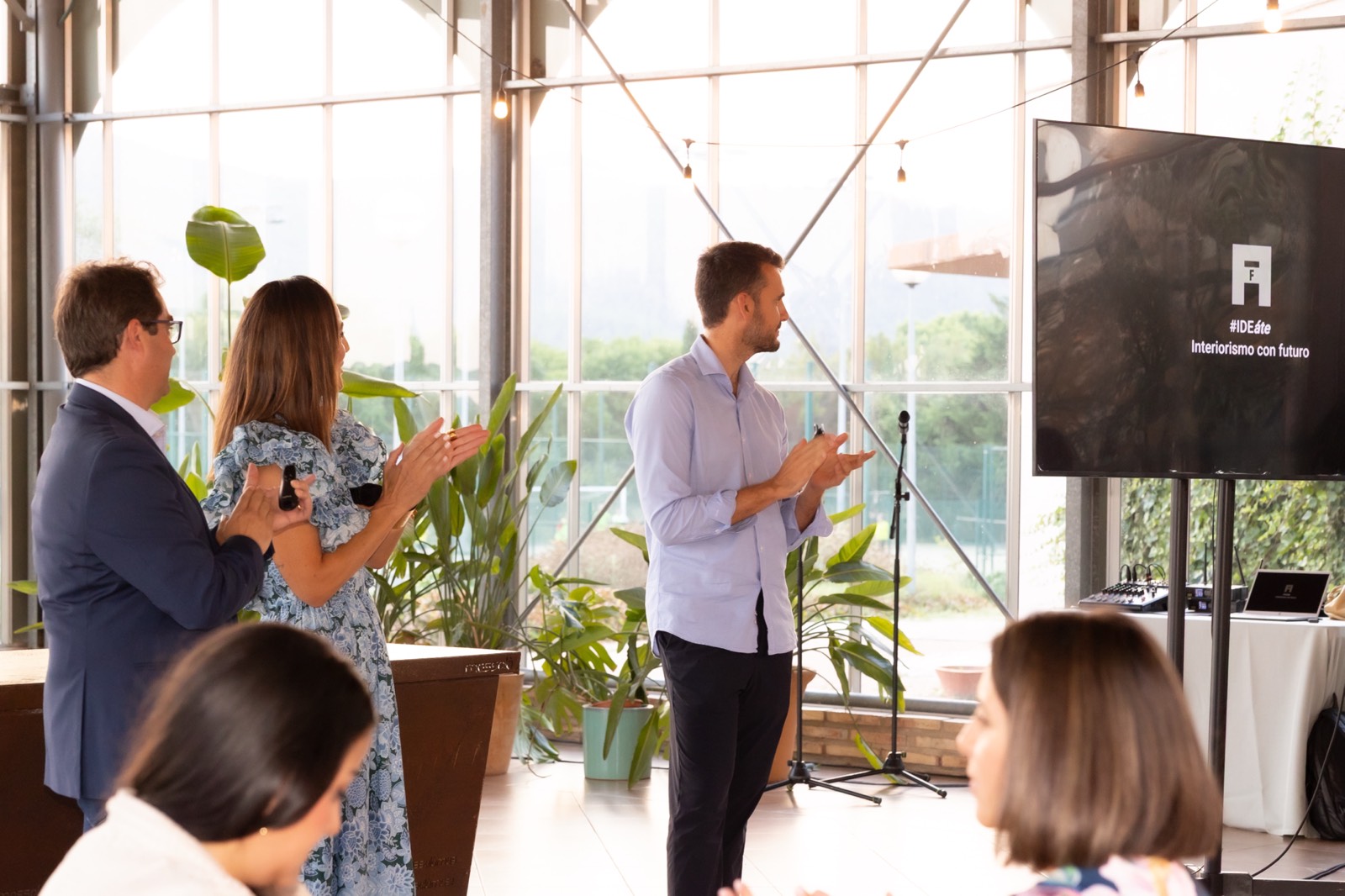 Presentación de evento profesional con oradores frente a una pantalla, rodeados de plantas en un espacio luminoso con grandes ventanales