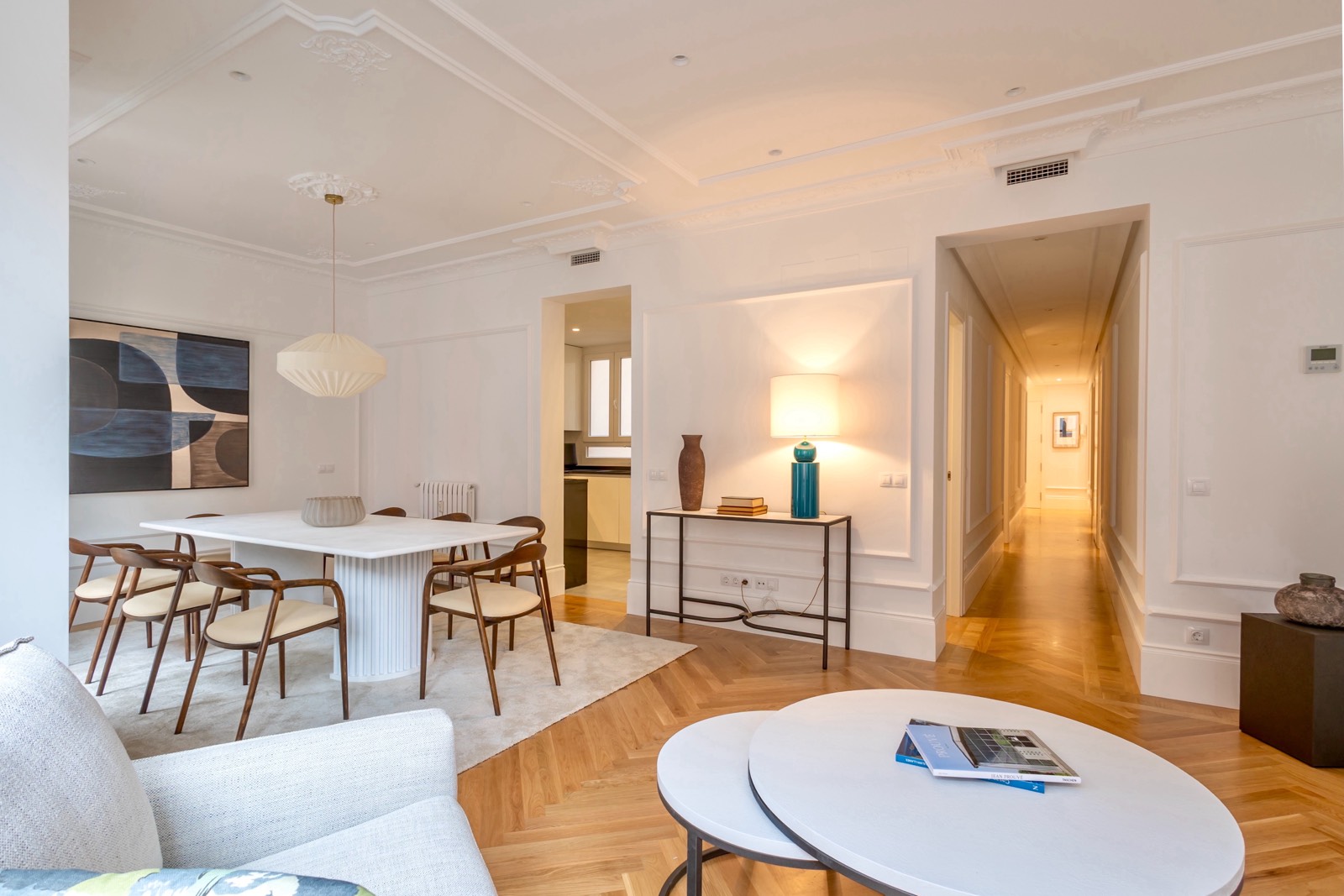 Elegante salón-comedor con mesa de mármol redonda, sillas de madera, lámpara colgante, suelo de parquet, techo alto con molduras y pasillo que conduce a otras habitaciones. Decoración en tonos claros y estilo contemporáneo.