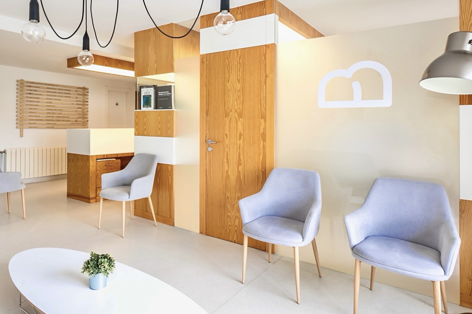 Sala de espera moderna con paredes de madera clara, sillas grises cómodas, iluminación colgante y una decoración minimalista. Se destaca una letra B luminosa en la pared.