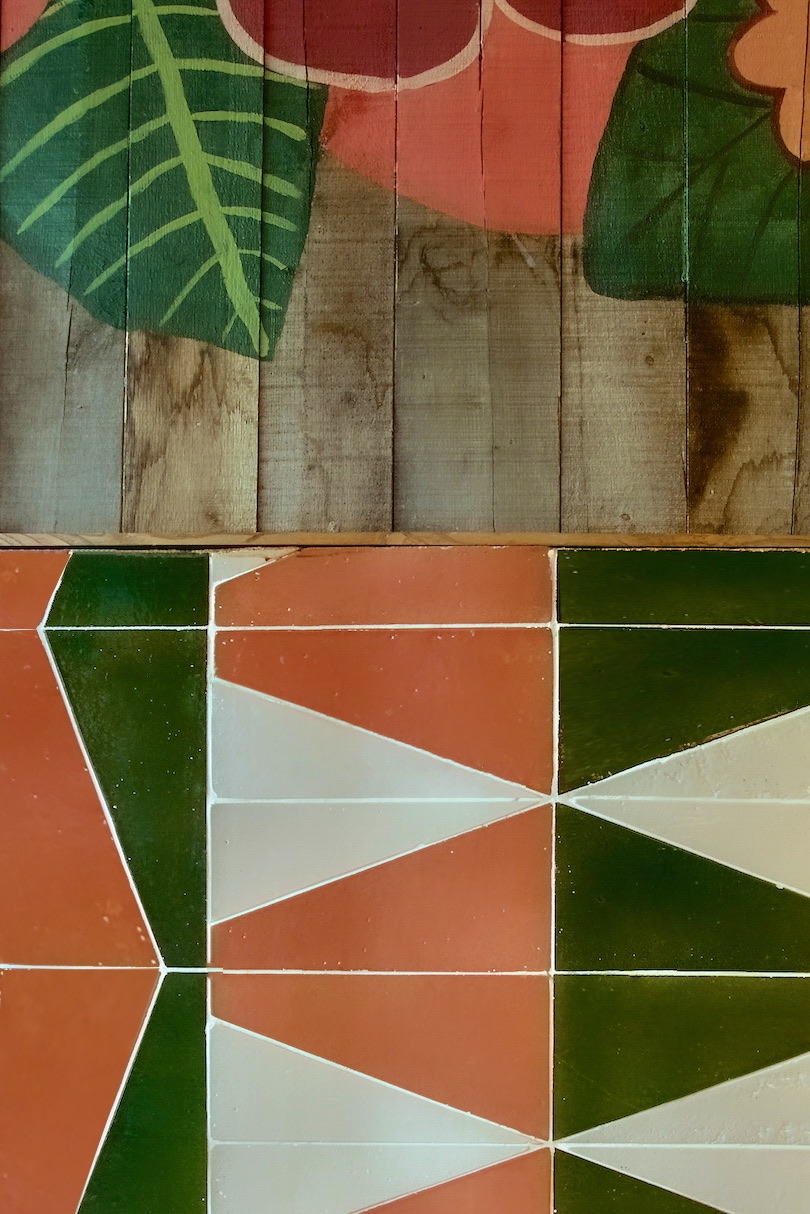 Diseño decorativo que combina elementos tropicales con hojas verdes en la parte superior y un patrón geométrico en la parte inferior con azulejos en colores verde, terracota y blanco formando diseños triangulares y rectangulares