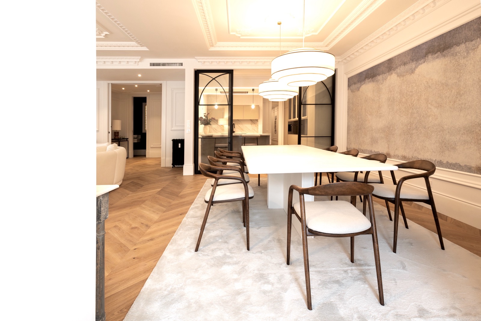 Comedor elegante y moderno con mesa blanca, sillas de madera, lámparas colgantes cilíndricas, piso de madera en espiga, y detalles arquitectónicos refinados como molduras en el techo y puertas arqueadas