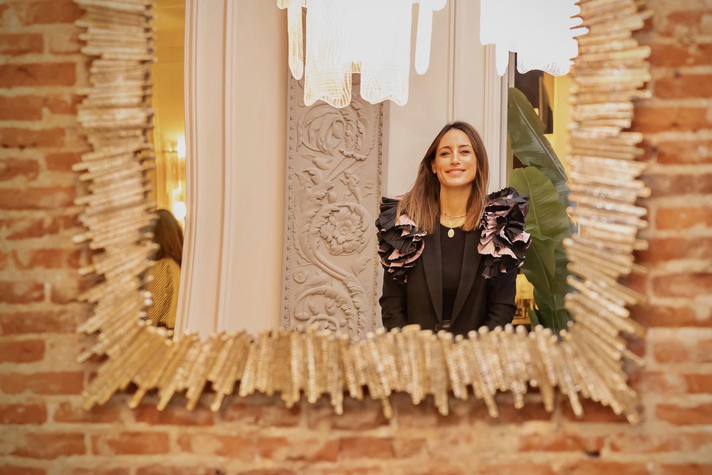 Imagen de una mujer sonriente reflejada en un espejo decorativo con marco dorado estilo sol. Ella viste una chaqueta negra con detalles voluminosos en los hombros. El fondo muestra una pared de ladrillo y elementos decorativos elegantes.