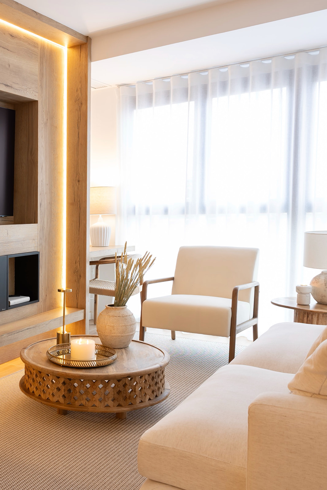 Sala de estar moderna con iluminación LED cálida, sofá blanco, sillón beige, mesa de centro de madera y ventanales grandes con cortinas blancas. Decoración minimalista y acogedora con tonos neutros.