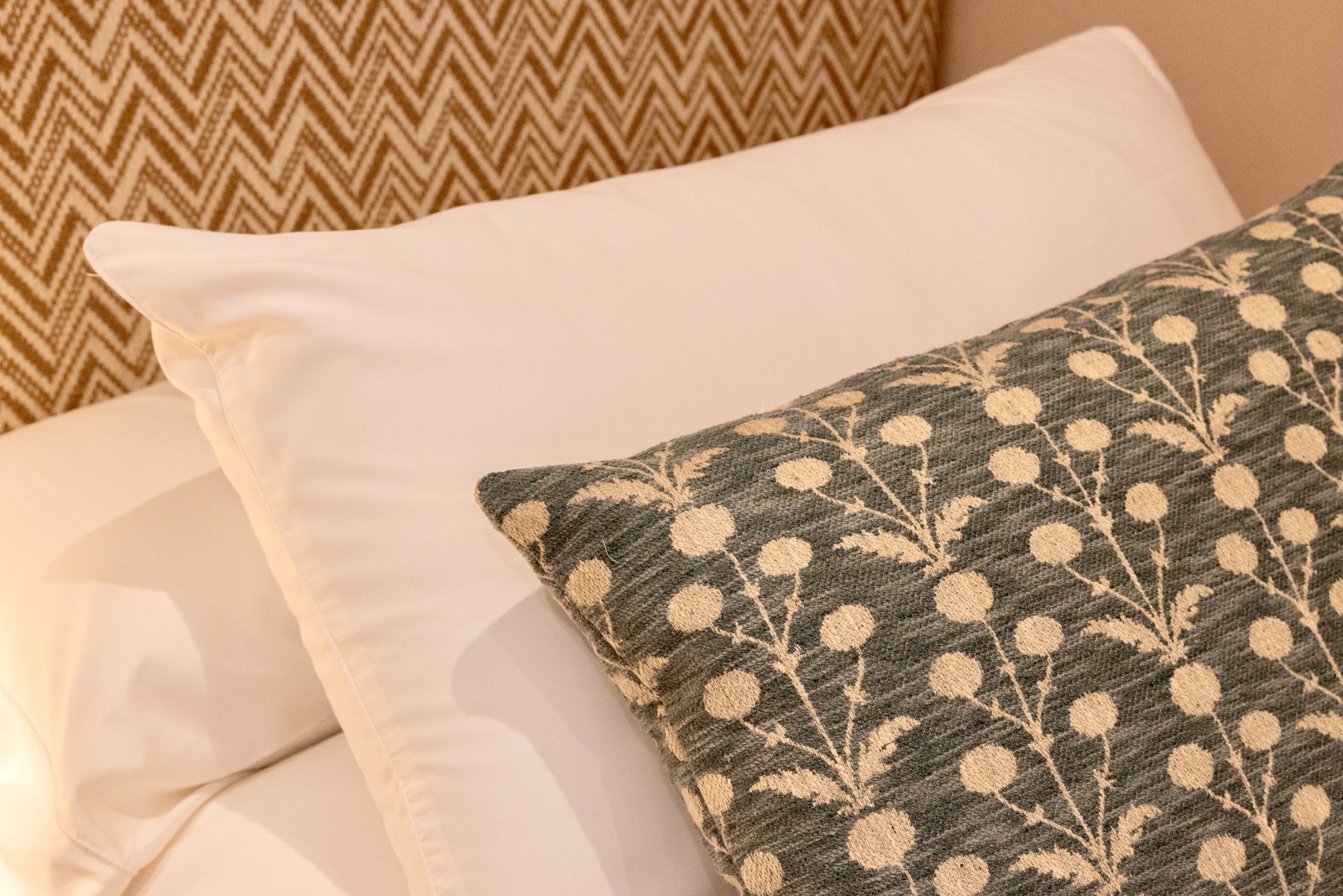 Primer plano de almohadas decorativas en una cama. Una almohada blanca y otra con un patrón floral gris y beige. El fondo muestra una tela con patrón de espiga.